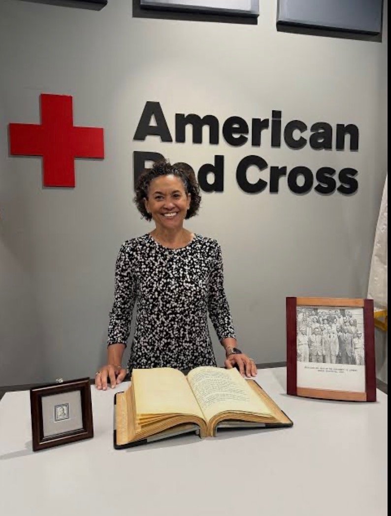 A woman standing behind a table with an open book and framed photographs, in front of a grey wall with the American Red Cross logo and text.