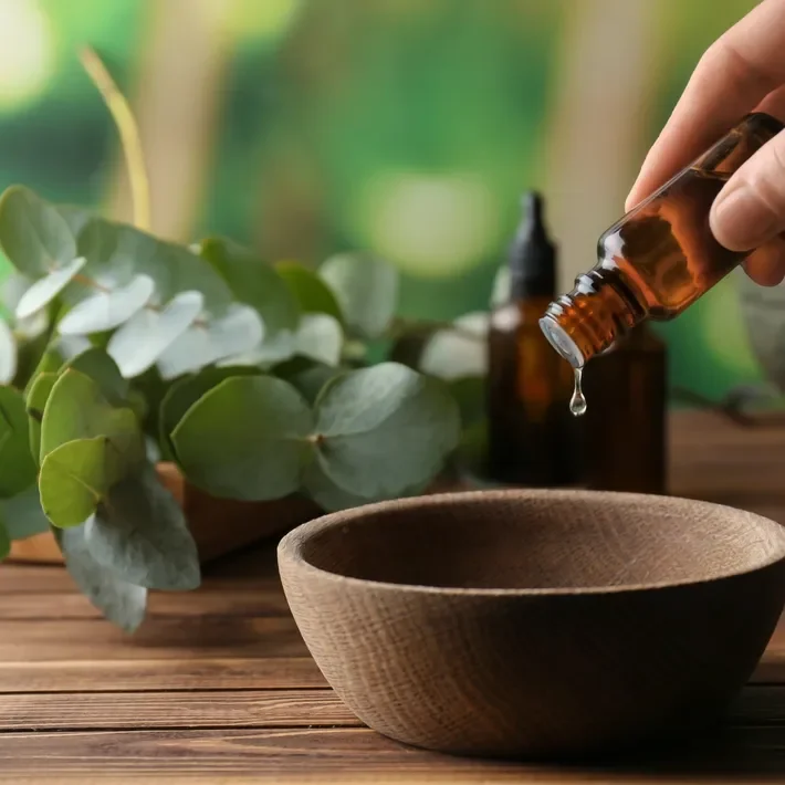 Dropper adding essential oil to a wooden bowl on a wooden surface, with eucalyptus leaves and an amber bottle in the background.