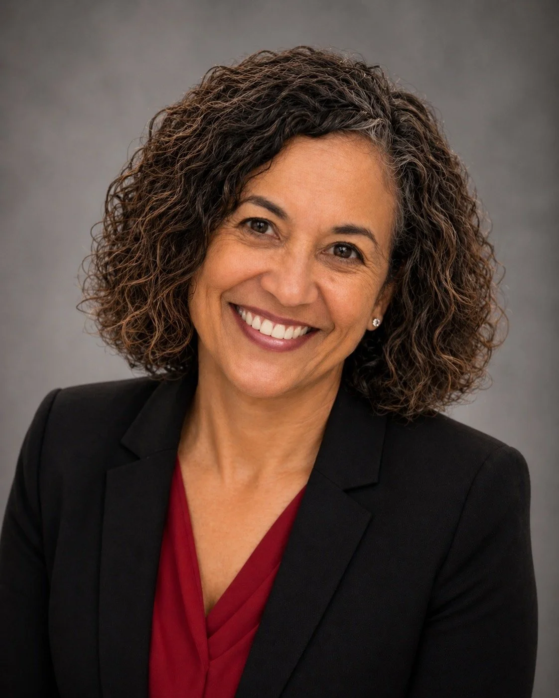 A professional woman with curly dark hair, smiling, wearing a black blazer over a red blouse, against a plain gray background.
