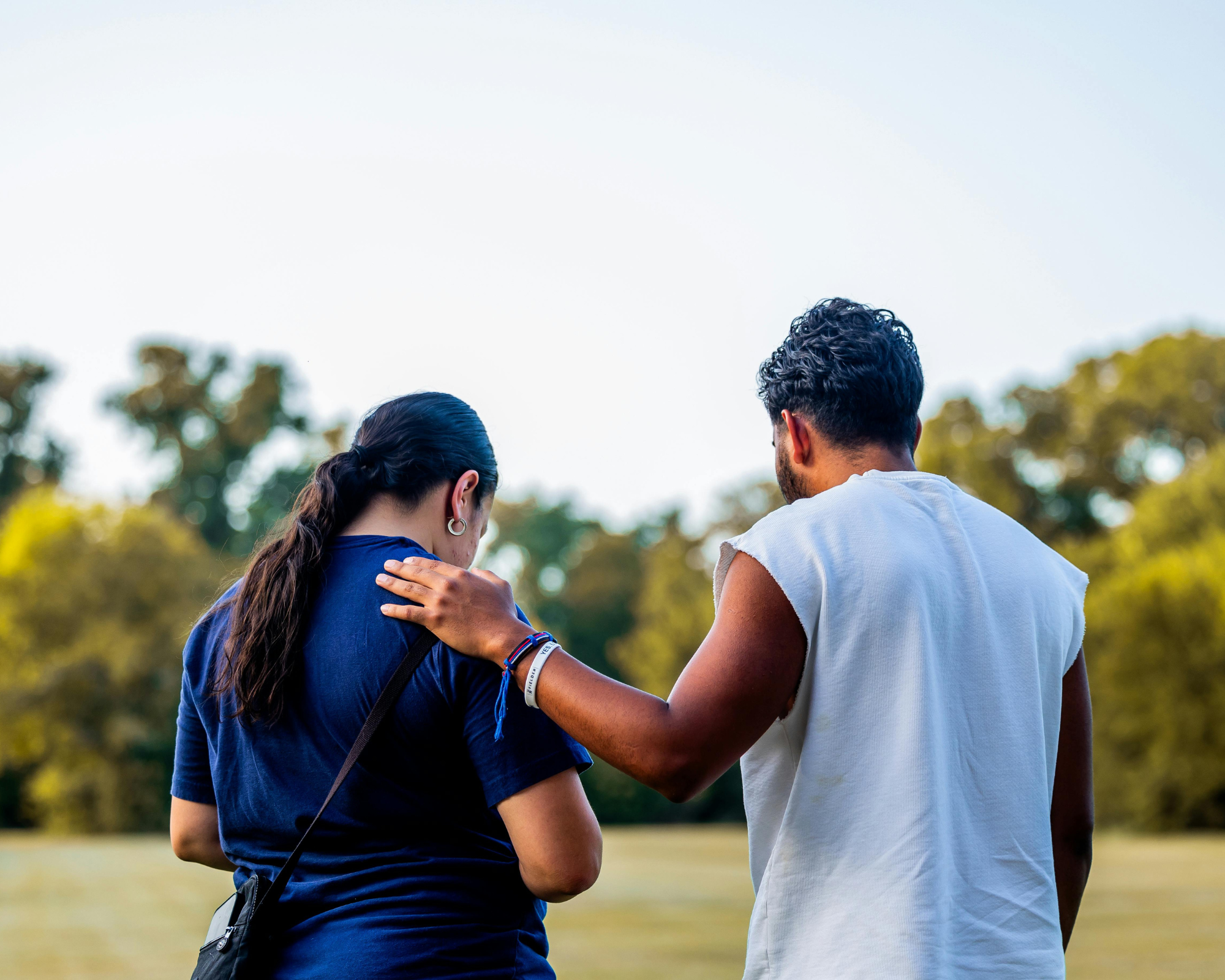 Two people embrace outdoors on a sunny day, with trees in the background. One has dark hair tied back, wearing earrings and a blue top. The other has curly hair, wearing a white sleeveless top. Their heads are close, with one person's arm around the other's shoulder.