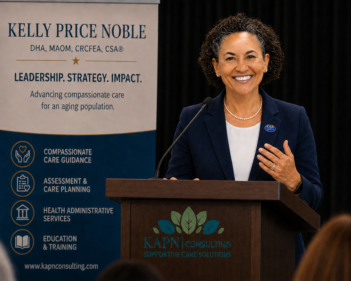 A woman in a navy blazer and pearl necklace standing at a podium, smiling, with a banner to her left that reads "Kelly Price Noble" and related health care information. There is a logo for Kapn Consulting on the podium.