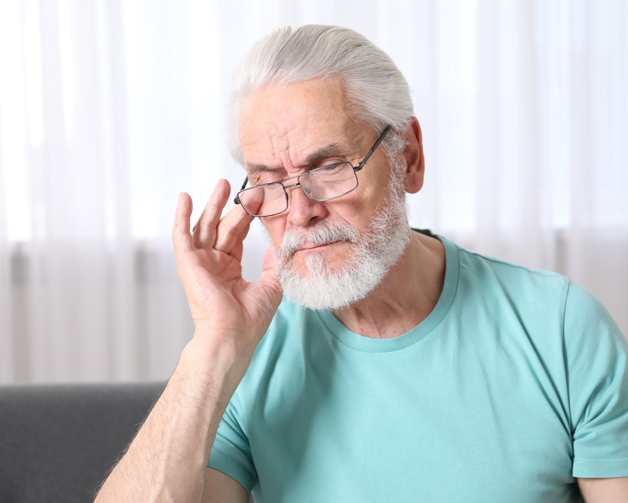 Older man with white hair and beard wearing glasses and a turquoise shirt, touching his forehead with his hand, appears to be in thought or concerned.