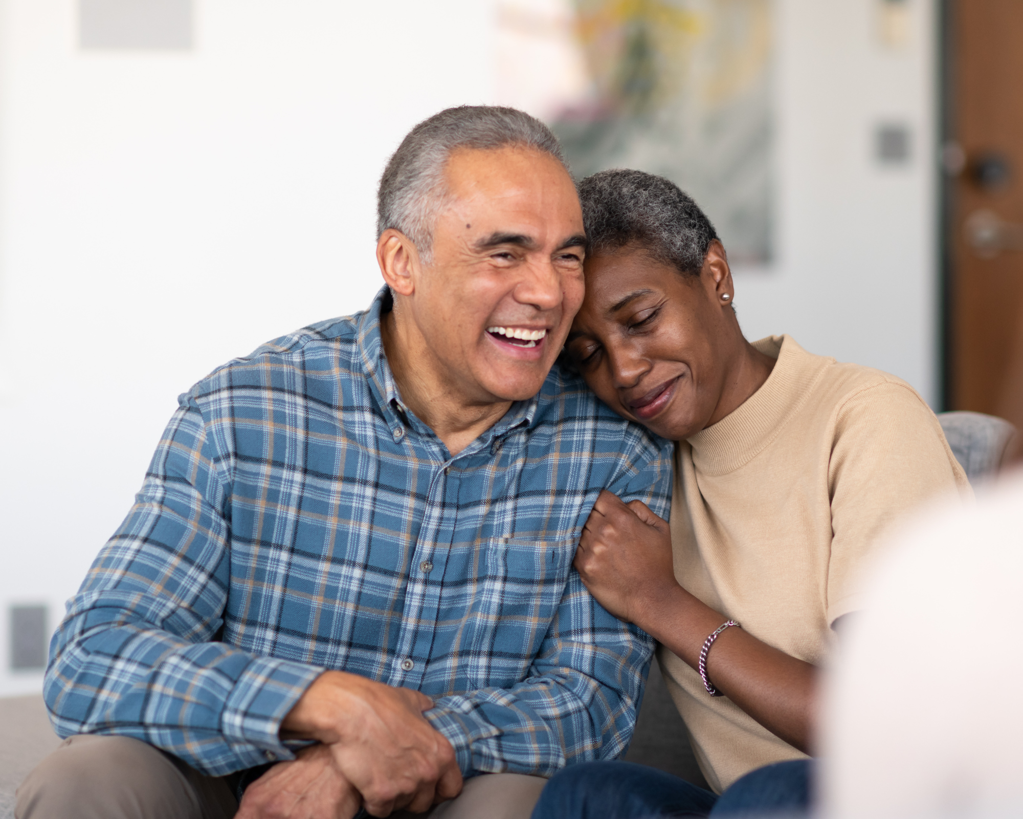 A smiling elderly man with gray hair and a checkered shirt sitting closely with a woman with short curly gray hair, wearing earrings and a light beige top, cuddling and expressing affection in a cozy indoor setting.