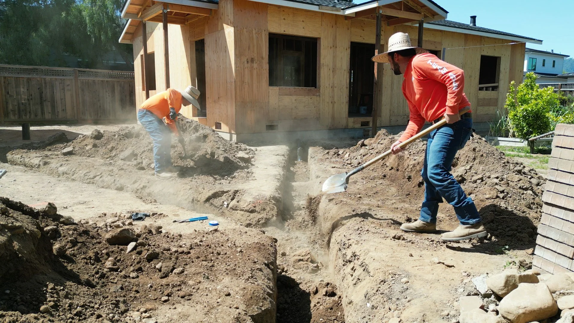 Two construction workers wearing wide-brimmed hats and long-sleeve shirts are digging a trench in the backyard of a house under construction. The house is covered in unfinished wood panels, and there are some tools and construction materials around.