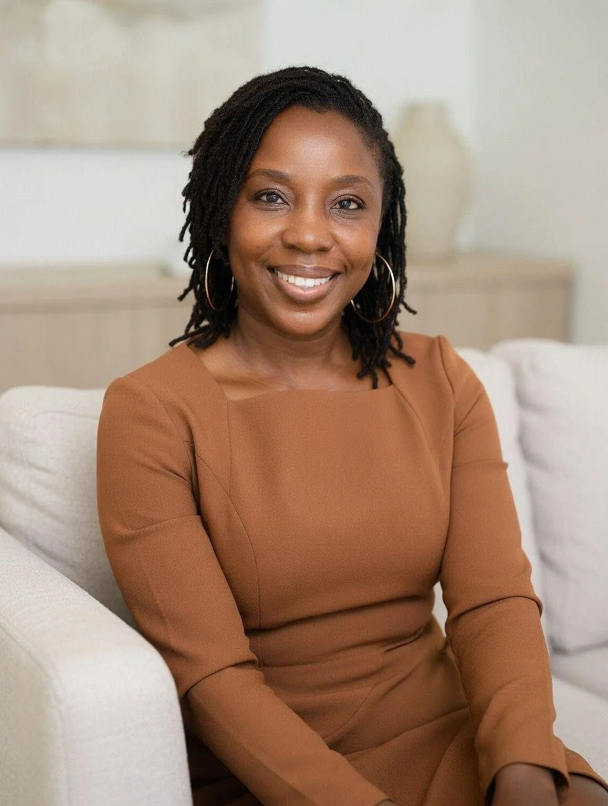 A woman with shoulder-length dreadlocks, wearing hoop earrings and a brown long-sleeved dress, smiling while sitting on a white couch in a bright room.