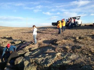 Group of people investigating rocky terrain near an open field with cars parked on the grass and a partly cloudy sky
