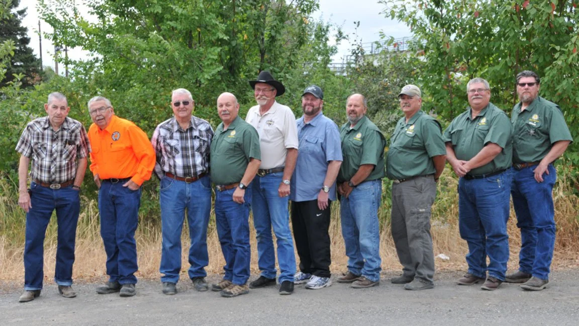 Group of eleven men standing outdoors in front of trees and bushes, some wearing green shirts with logos, one in an orange shirt, and one in a white shirt and cowboy hat.
