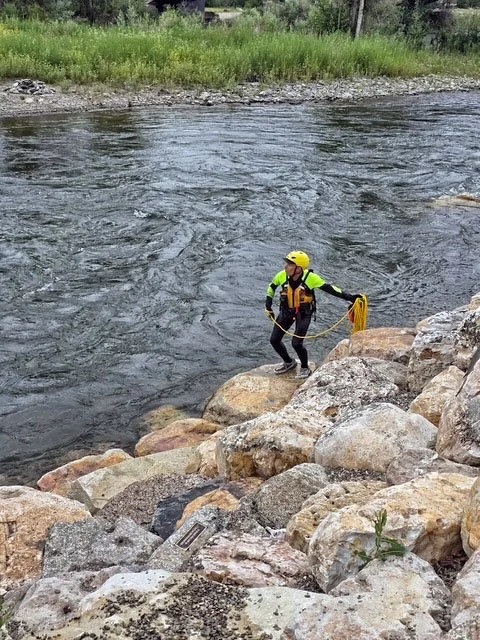Swift Water Rescue Training - A person wearing a yellow helmet, a bright yellow and black jacket, and a life vest holding a rope while standing on rocks near a river.
