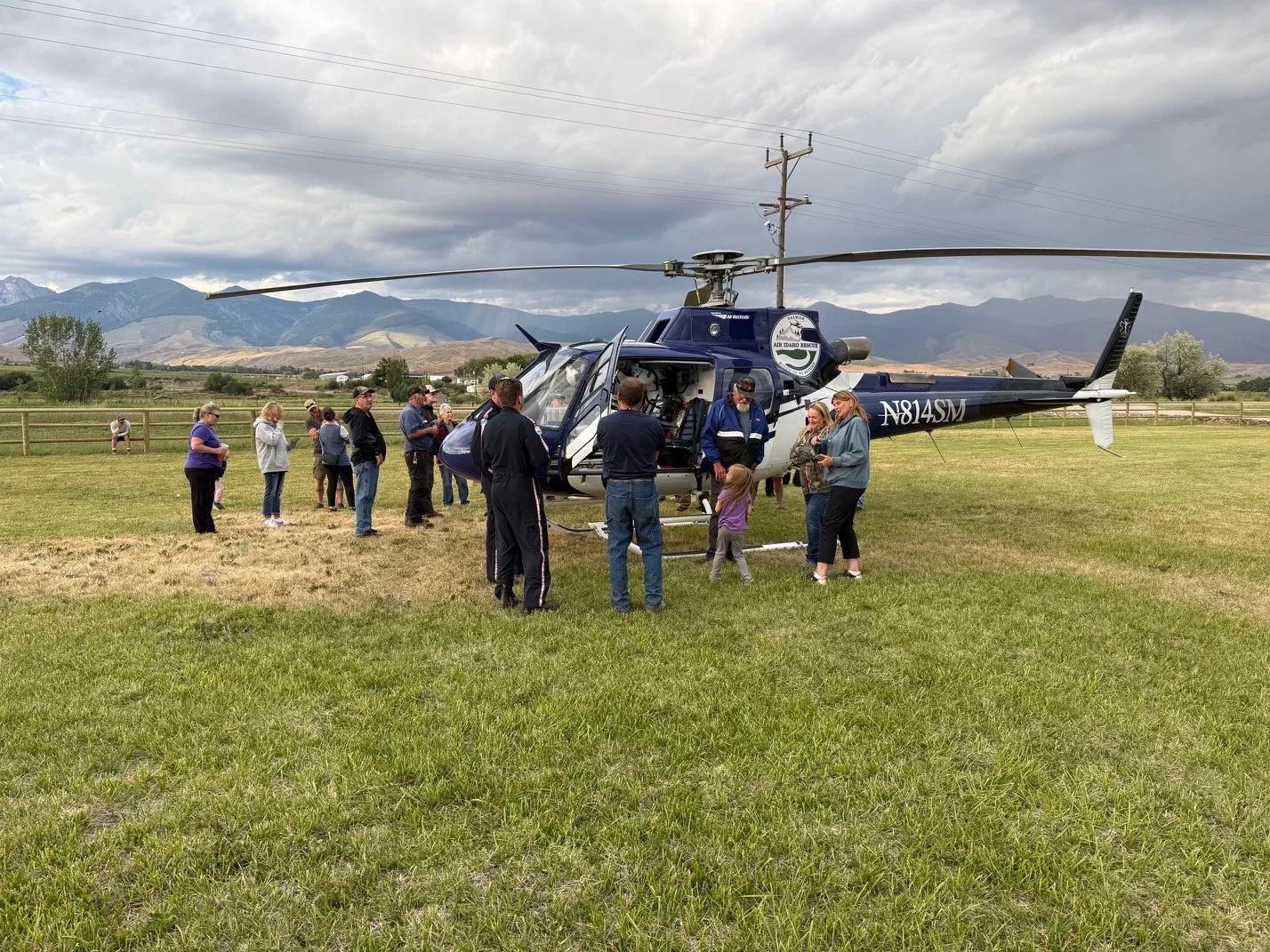 An Air Idaho Rescue helicopter on a grassy field with a group of people gathered around it, some talking and others looking inside. There are mountains in the background and electrical poles overhead under a cloudy sky.