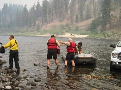 Four rescue workers in life vests assist a person near a boat on a riverbank, with a vehicle parked nearby.