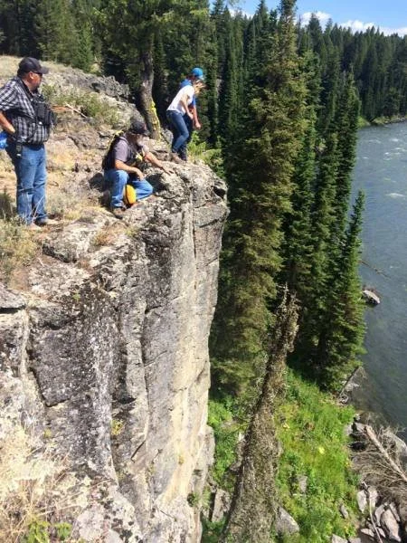 Four people standing and crouching on a rocky cliff edge above a river, surrounded by trees.