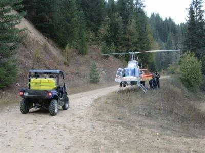 A helicopter landing on a dirt road, with a golf cart nearby and people helping with the landing in a forested area.