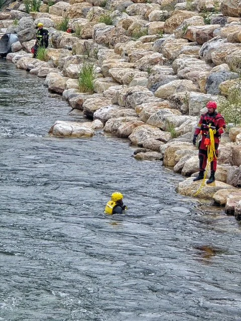 Swift Water Rescue Training - Rescue workers in safety gear perform a water rescue in a river, with one person in the water and others on the rocky riverbank.