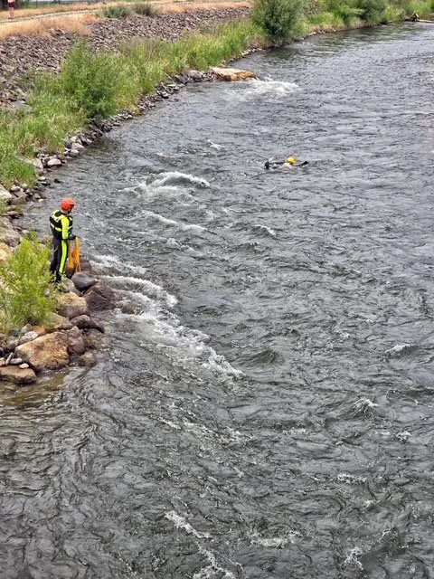Swift Water Rescue Training - Person in a wetsuit and helmet swimming in a river with a yellow helmet nearby, another person in safety gear standing on the rocky riverbank, lush greenery on the riverbank.