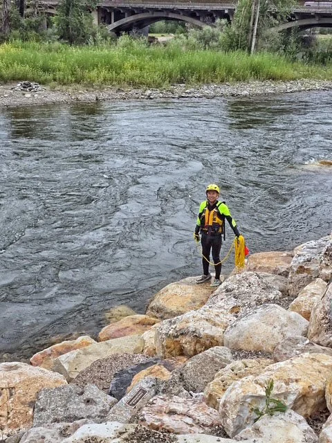 Swift Water Rescue Training - Person in safety gear standing on rocks near a river, holding a rope, with a bridge and greenery in the background.