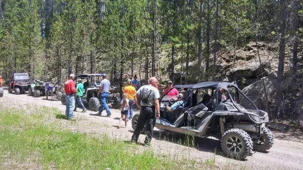 Group of people gathered around off-road vehicles parked on a dirt trail in a forested area.