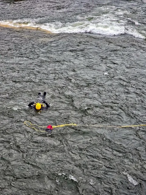 Swift Water Rescue Training - Person swimming in a dark river, wearing a yellow helmet and a black wetsuit, with a yellow rope attached to something outside the frame.