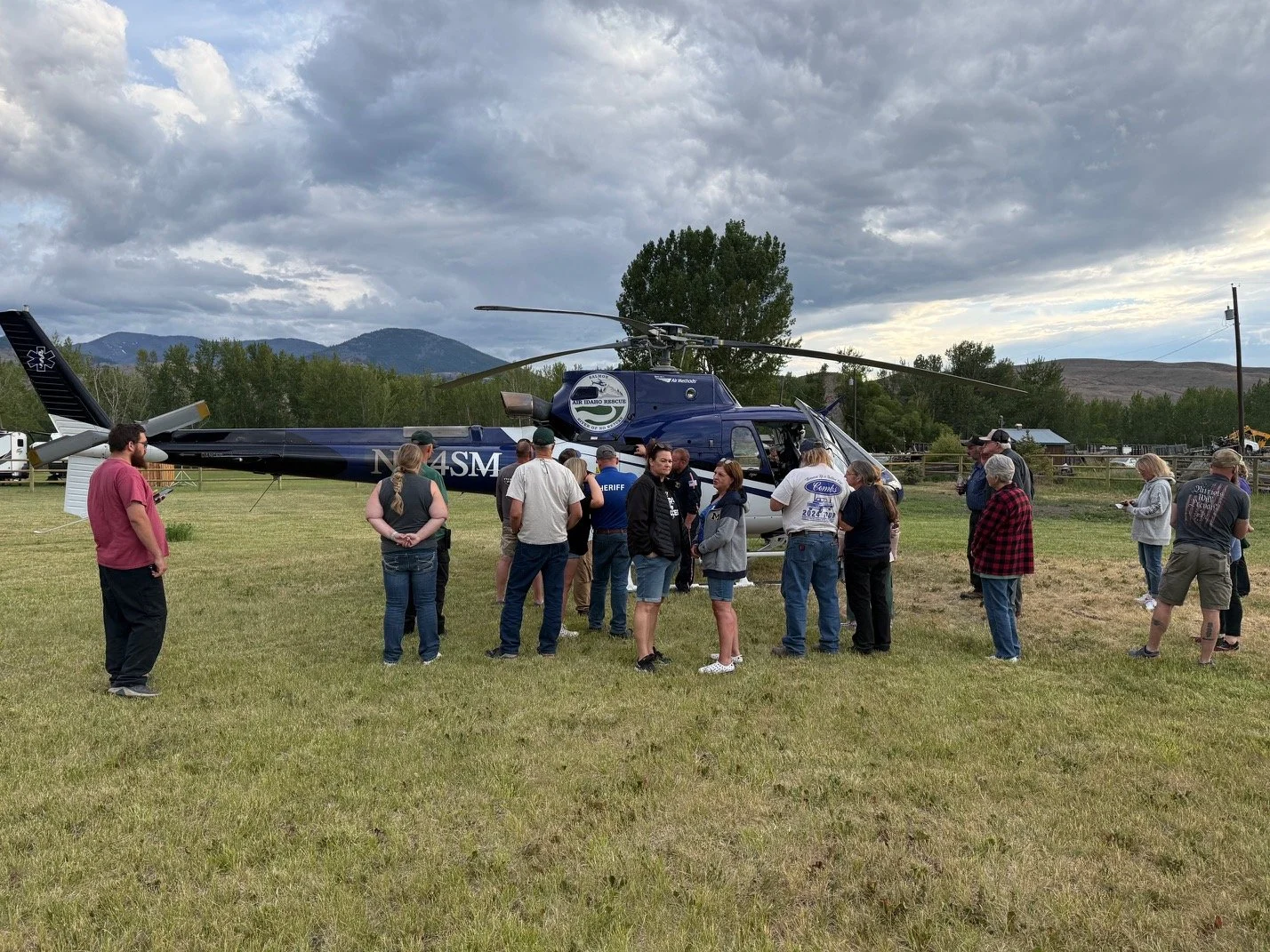 A group of people gathered around an Air Idaho Rescue rescue helicopter on a grassy field, with mountains and cloudy sky in the background.