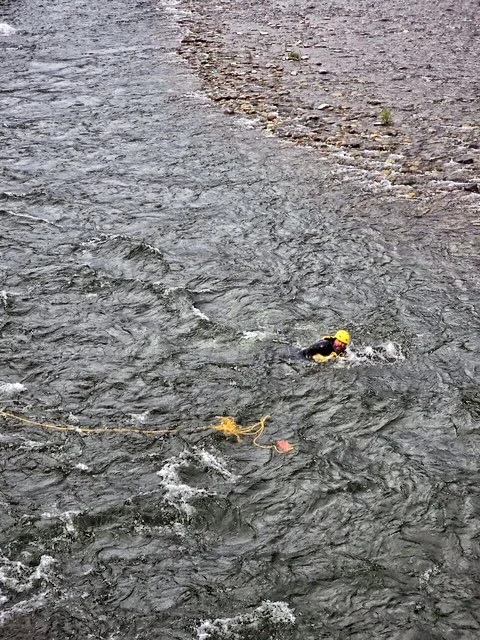 Swift Water Rescue Training - A person swimming in a rocky river wearing a yellow helmet and dark swimwear, holding onto a rope