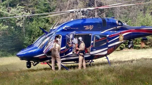 A blue helicopter with red and white accents on a grassy field, with three people near the open door.