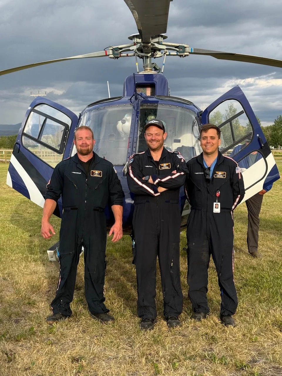 Three male pilots in black uniforms standing in front of an Air Idaho Rescue helicopter on a grassy field, with a cloudy sky above.