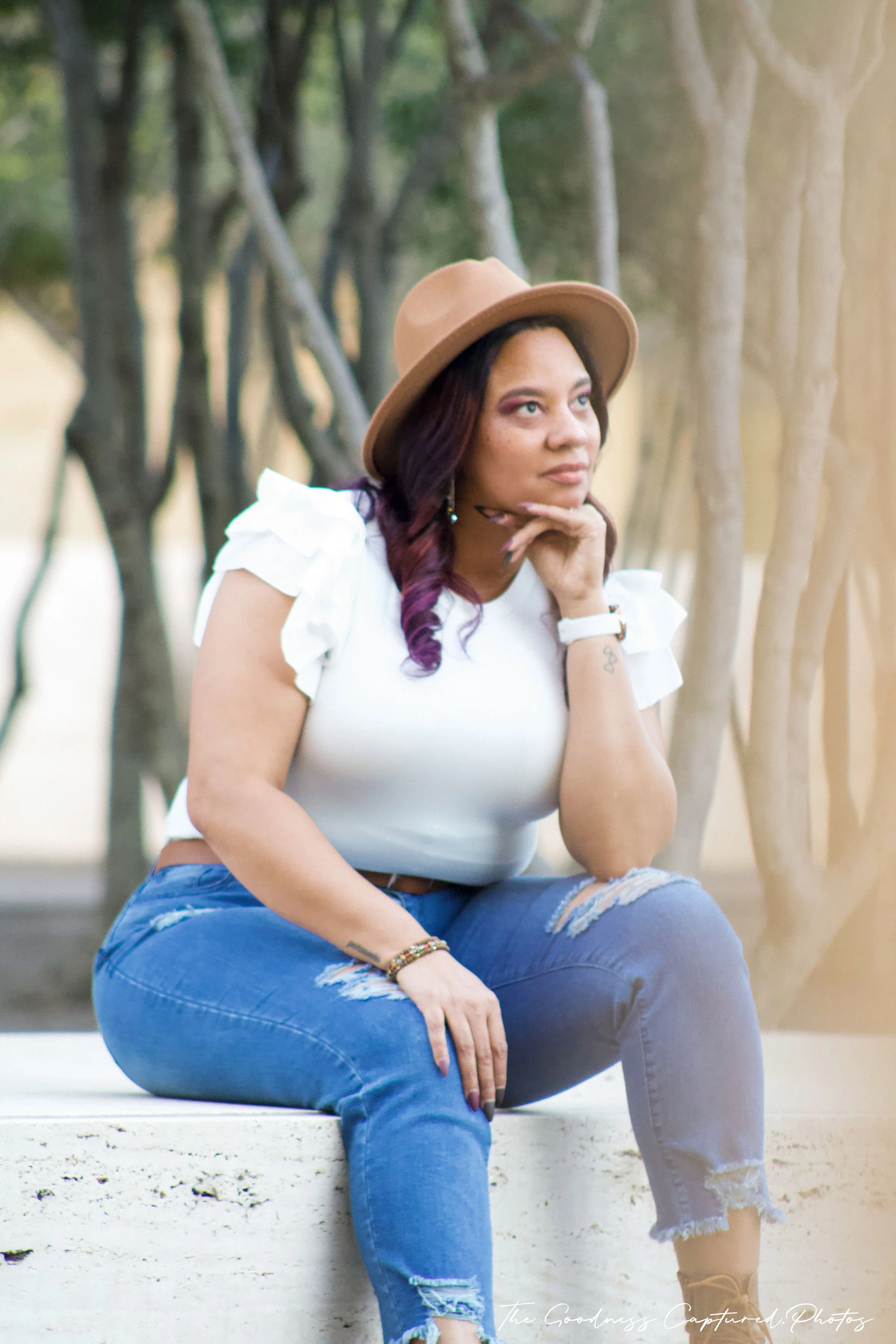 A woman with red and purple hair wearing a tan hat, white ruffled sleeve top, and ripped jeans, sitting on a white ledge outdoors with trees in the background.