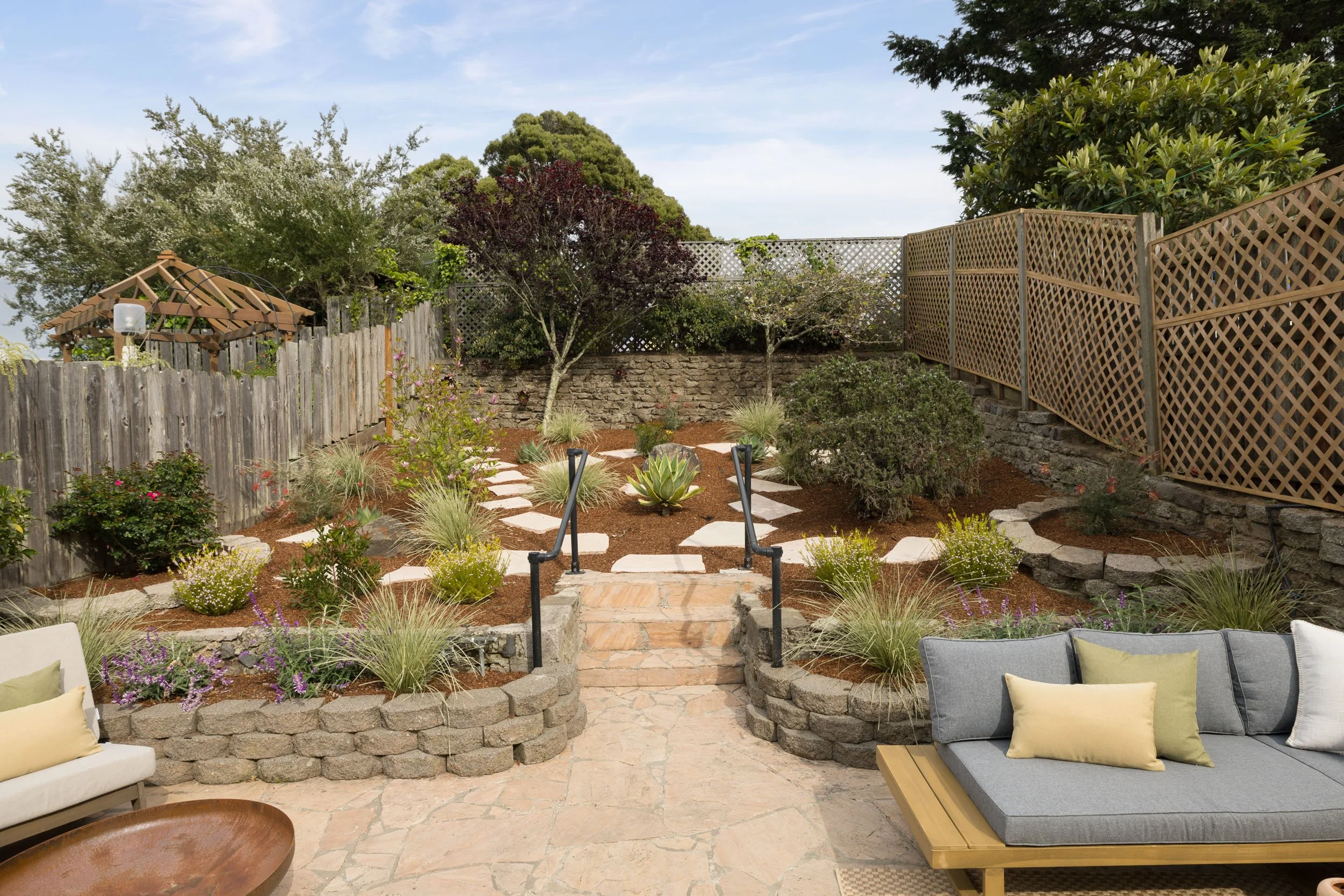 Backyard garden with stone pathway, various shrubs, and trees, enclosed by wooden fencing, with outdoor seating in the foreground.
