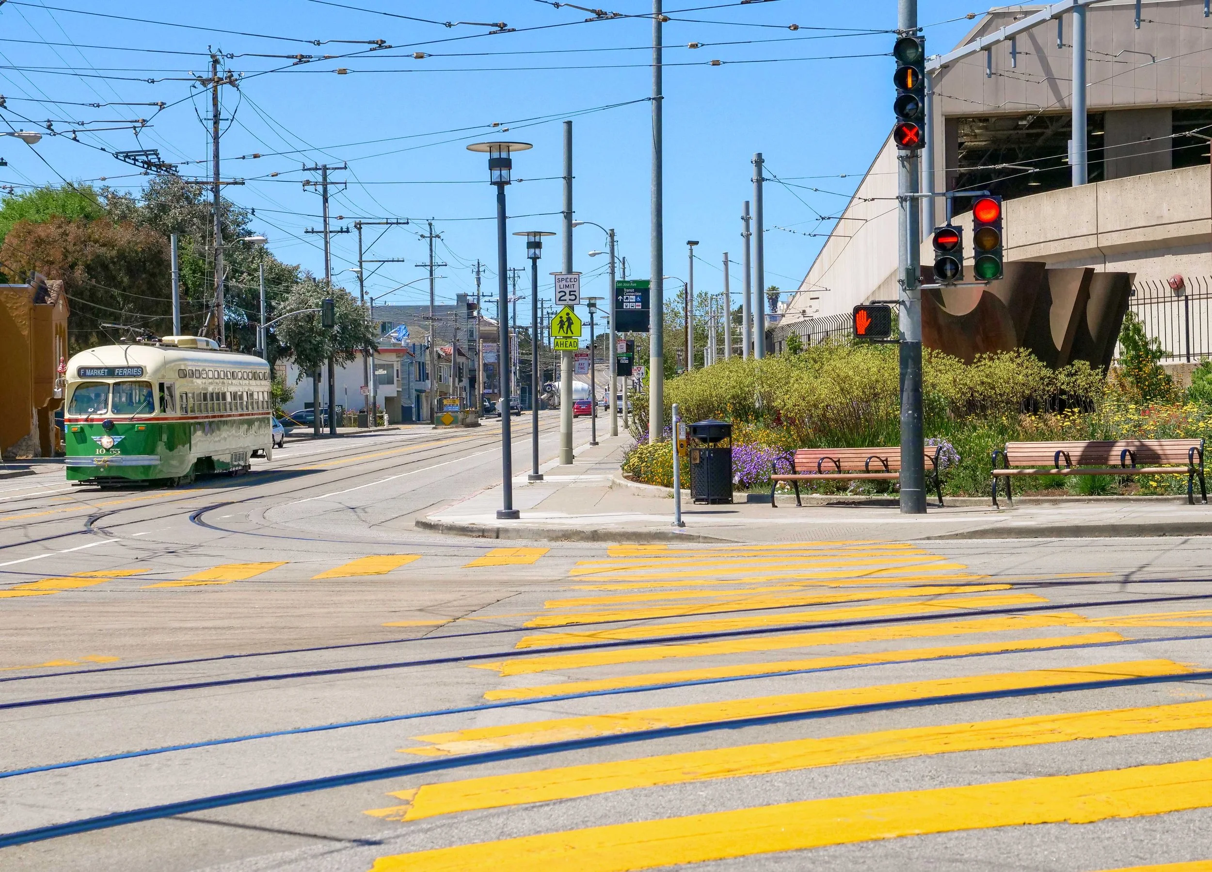 City street intersection with yellow crosswalk, traffic lights showing red and green, a green and white trolley bus on the tracks, benches, and flowers in a landscaped area.