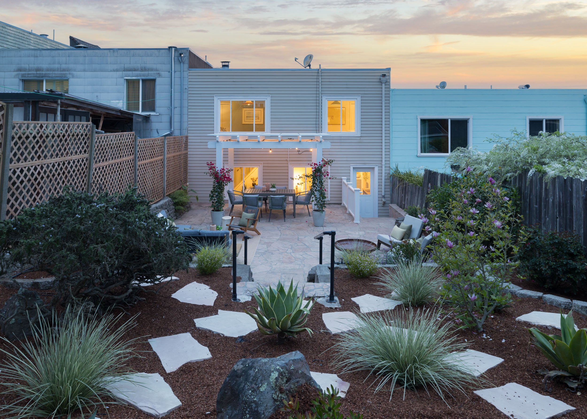A backyard patio area at dusk with outdoor seating, plants, and a two-story house with lit windows in the background.