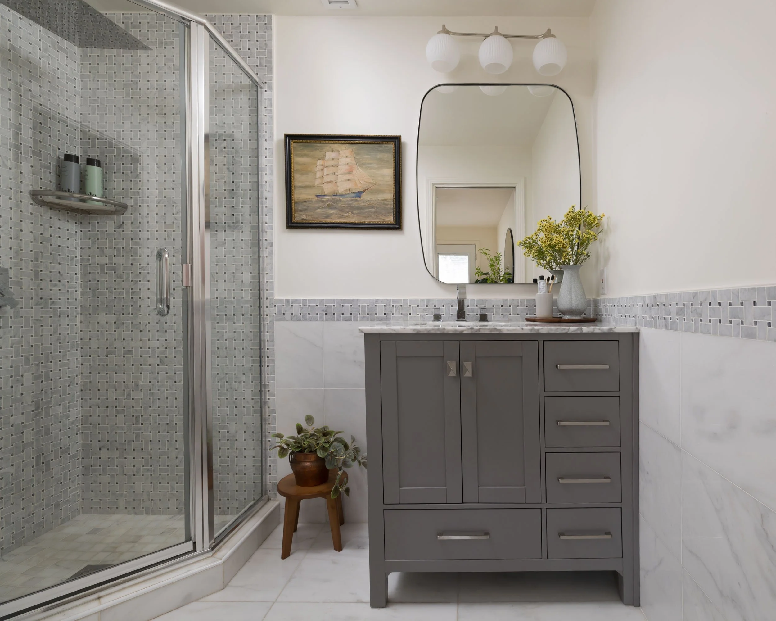 Modern bathroom with a walk-in shower, gray vanity with drawers, a large mirror, a framed painting of a sailing ship, a vase with flowers, and a potted plant on a wooden stool.