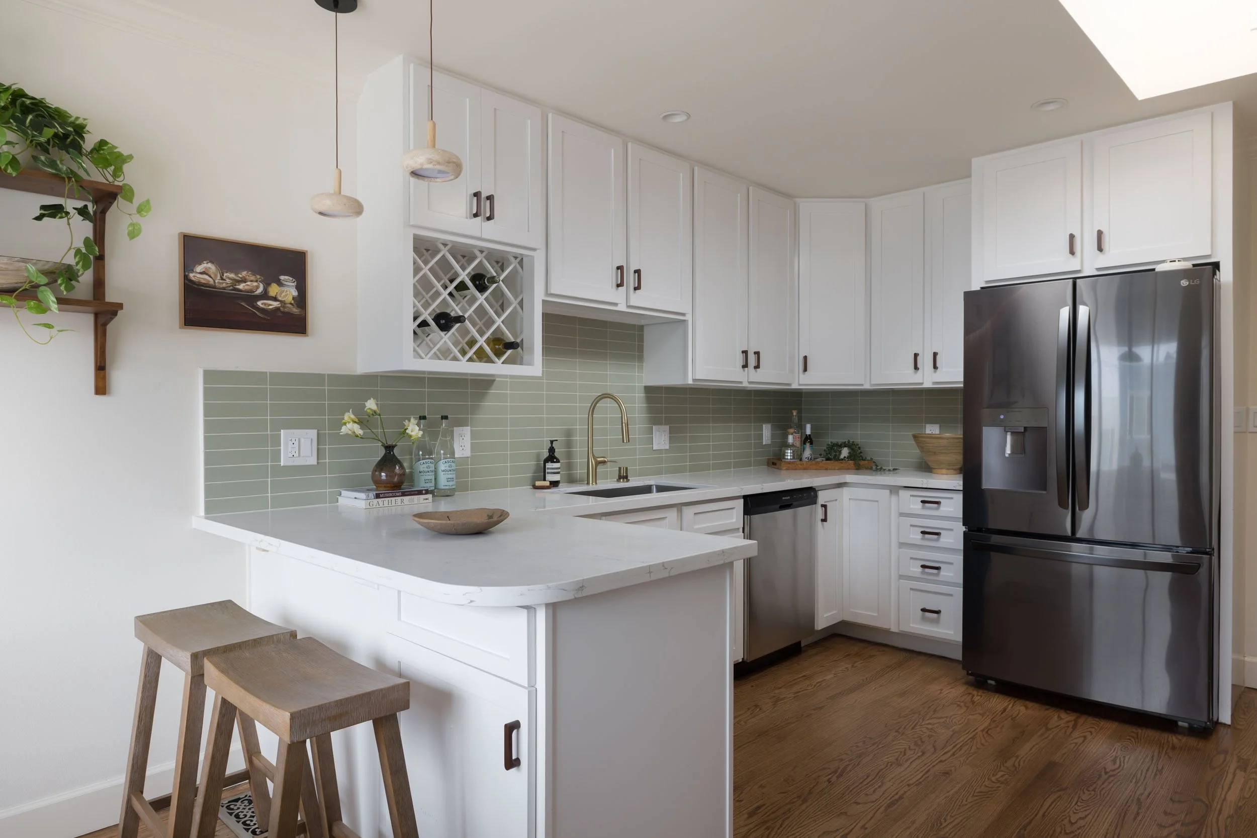 Modern kitchen with white cabinets, green tile backsplash, stainless steel refrigerator, black dishwasher, pendant lights, and wooden bar stools.