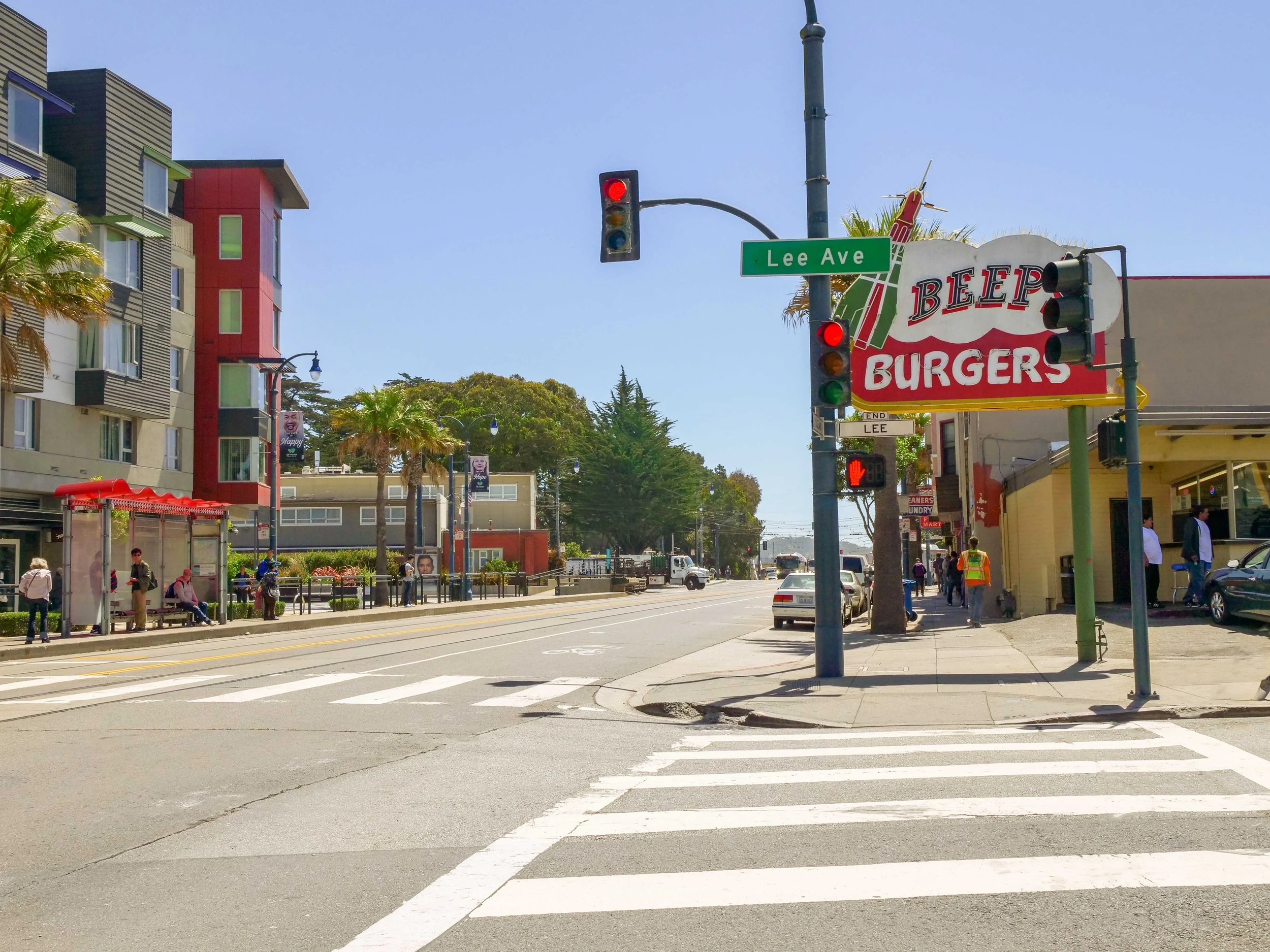 A city street corner with a green street sign reading 'Lee Ave', traffic lights, a bus stop with benches, palm trees, pedestrians, some waiting at the bus stop, and a red and white sign for 'Bleep Burgers' on a building.
