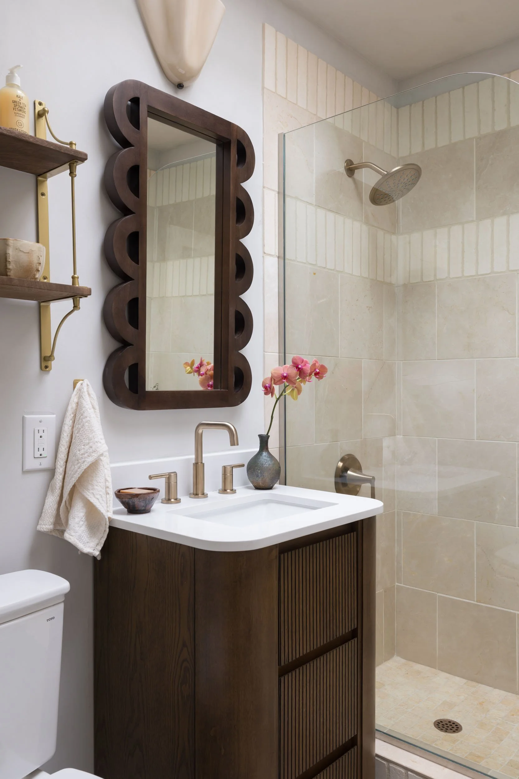 A modern bathroom featuring a wooden framed mirror, a white vanity with a gold faucet, a vase with pink orchids, a shower with beige tiles, and a clear glass shower door.