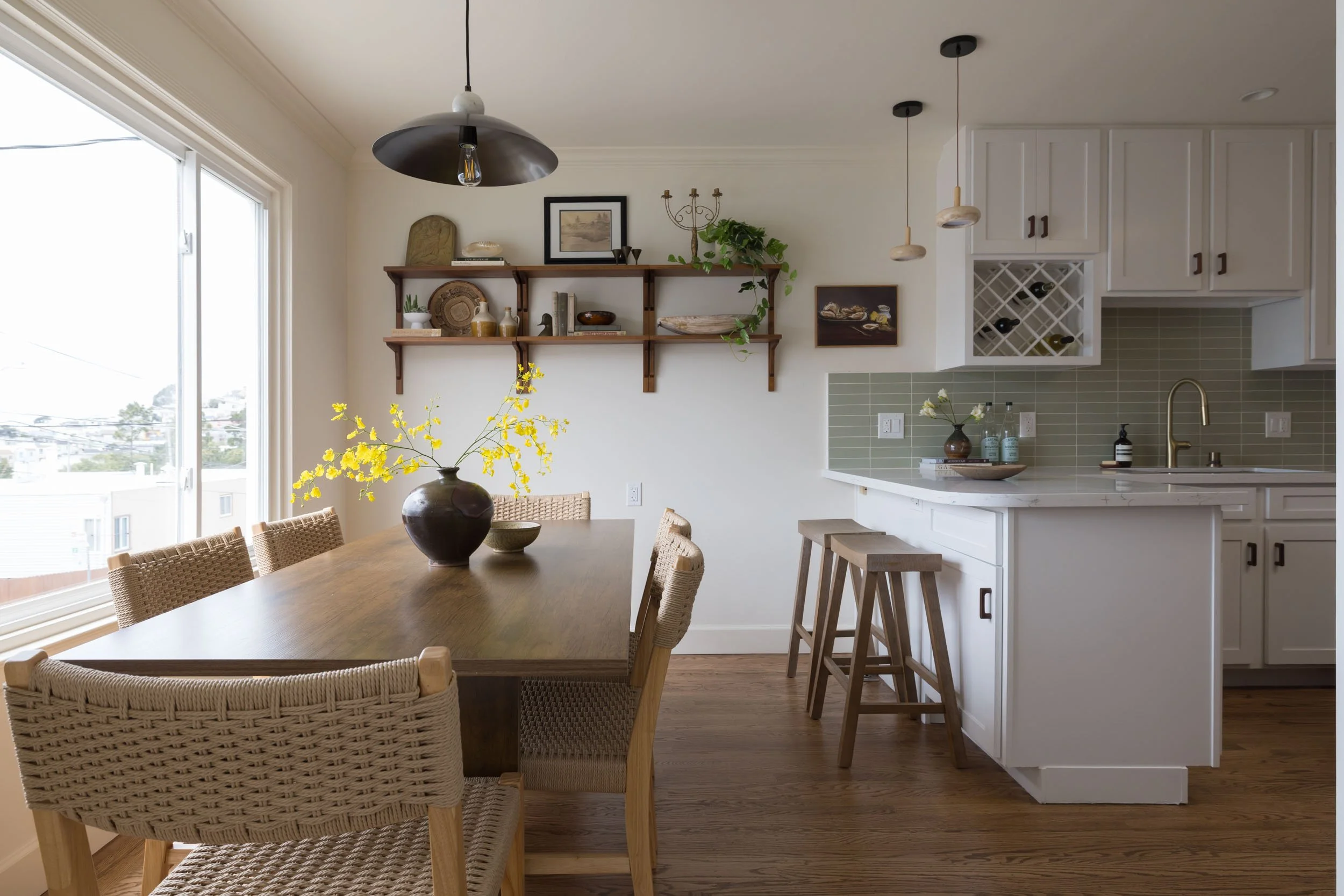 A dining area with a wooden table, six woven chairs, and a vase with yellow flowers. Adjacent to the dining area is a kitchen with white cabinets, green tile backsplash, and wooden barstools. There are wall shelves with decor and a large sliding glas