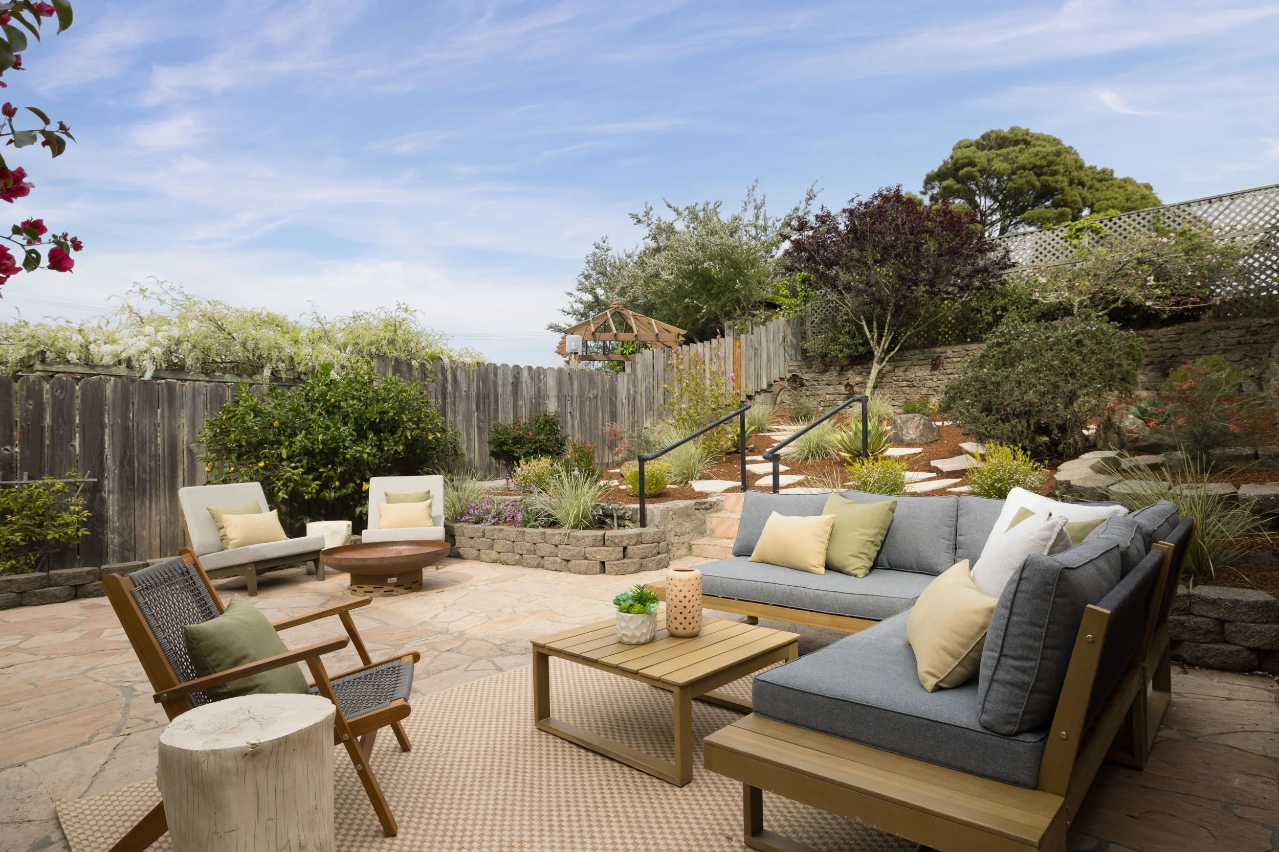 Outdoor patio with stone flooring, cushioned seating, and a garden with plants, rocks, and a wooden fence in the background.