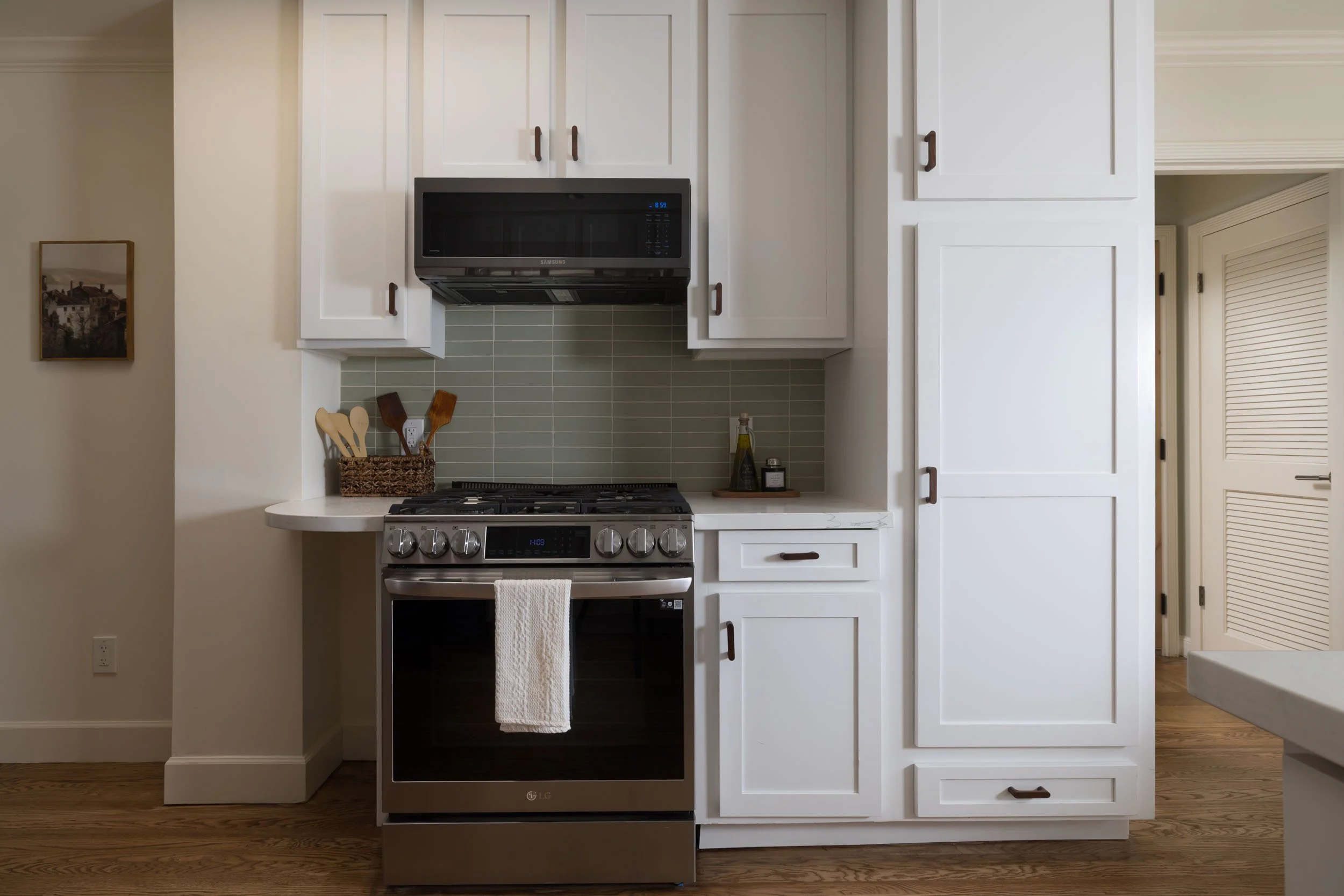 A modern kitchen with white cabinets, a stainless steel gas stove with an oven, a microwave above, and a light green tile backsplash. A towel hangs from the oven handle, and a basket with wooden cooking utensils is on the countertop. There are decora