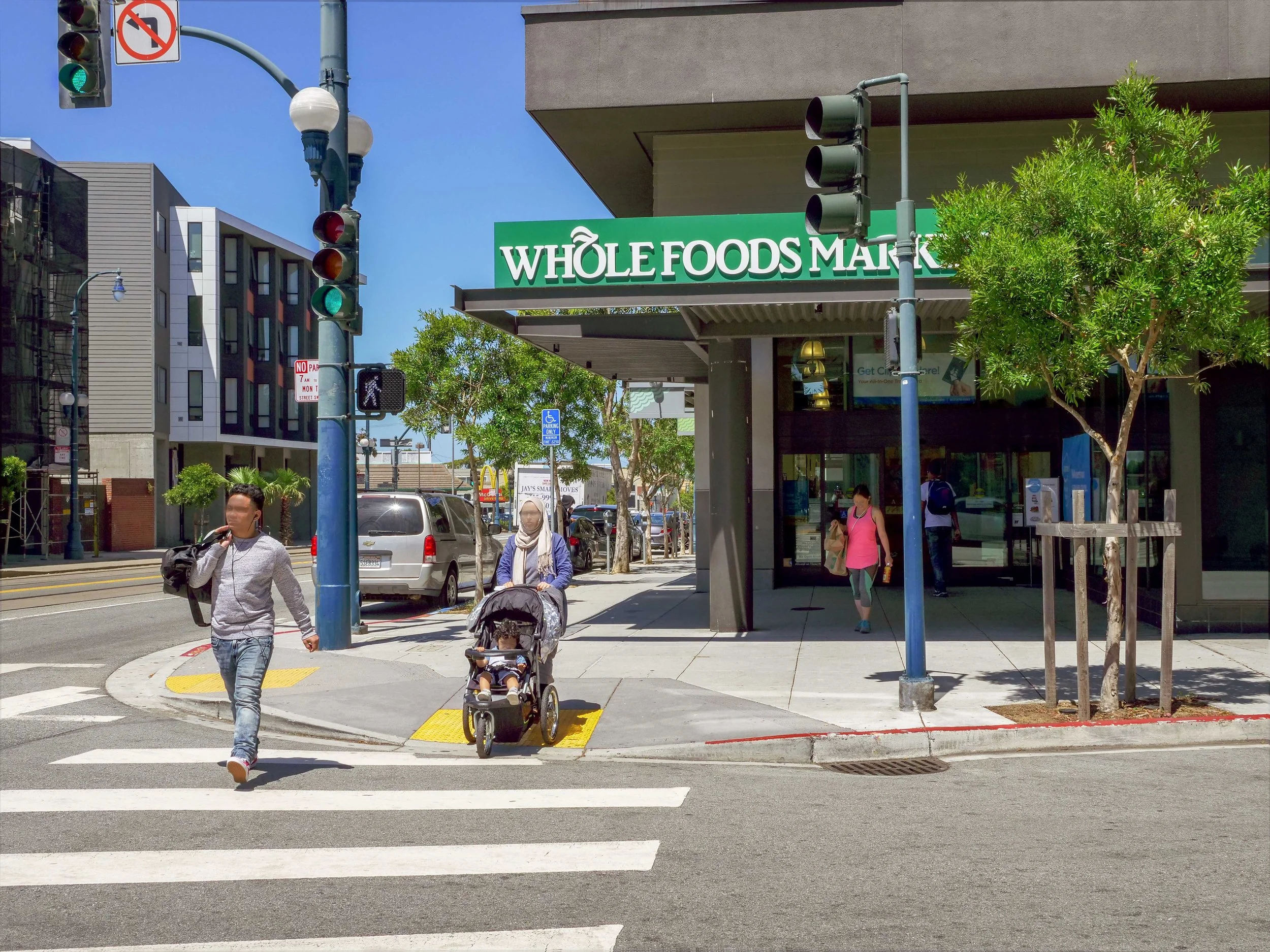 People crossing the street in front of a Whole Foods Market, with trees, cars, and a traffic light in the scene on a sunny day.