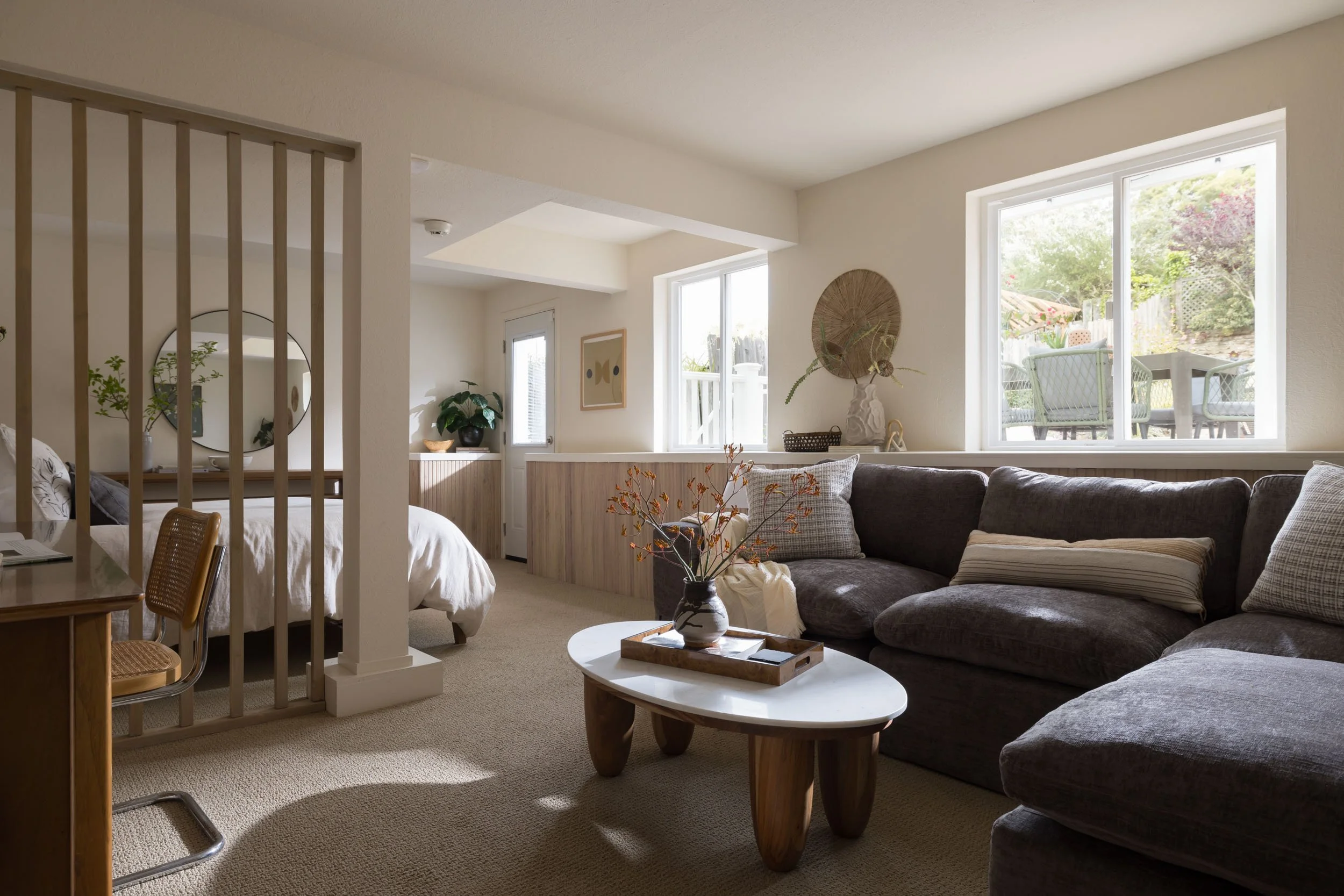 A cozy living room with large windows, a dark gray sectional sofa with pillows, a white marble coffee table with a vase and decorative branches, and a bedroom visible in the background separated by wooden slats.