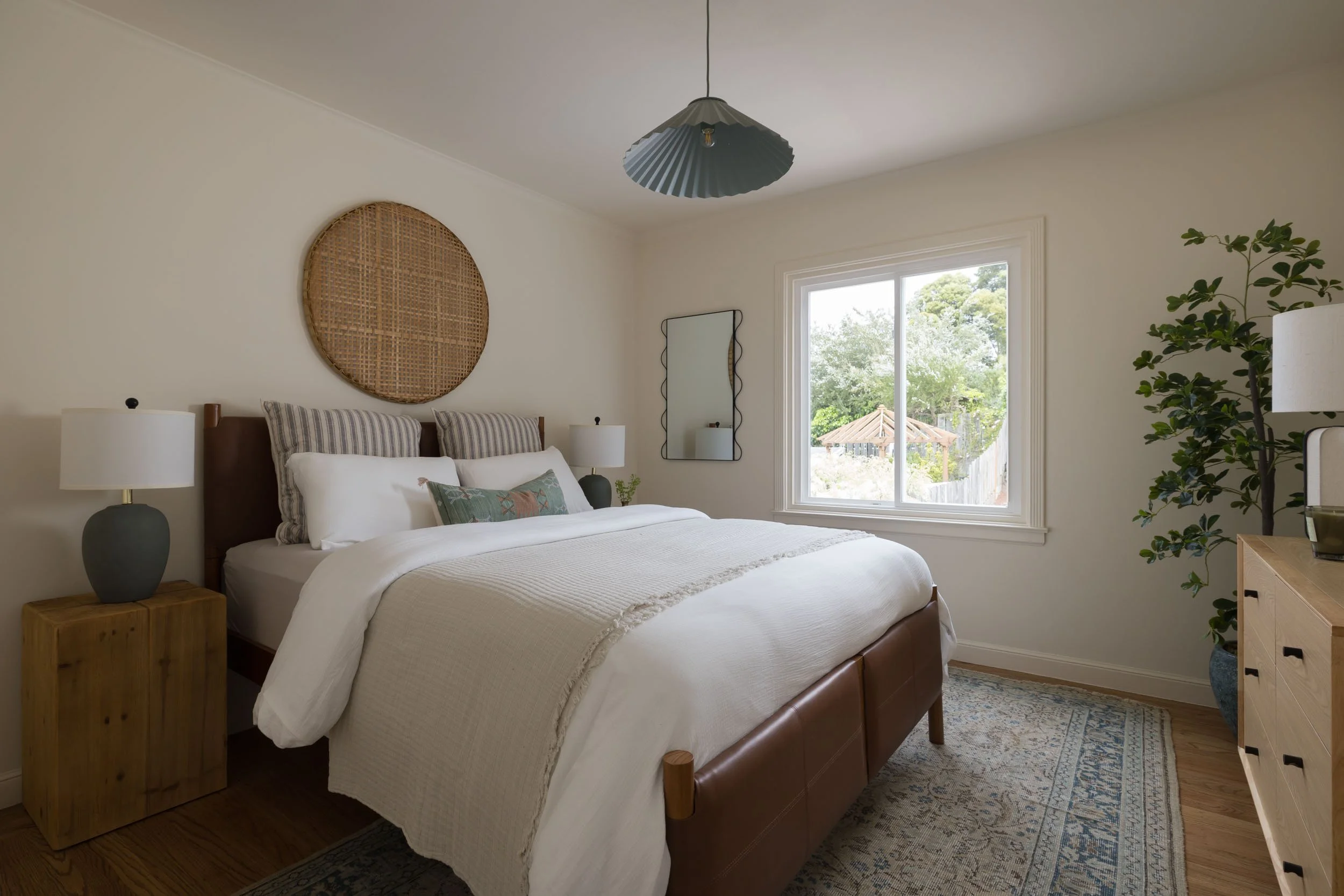 A cozy bedroom with a large bed, white bedding, striped and decorative pillows, and nightstands with lamps. A window shows trees and a gazebo outside, and there is a plant in a pot. The room has a beige rug and simple decor, including a round woven w