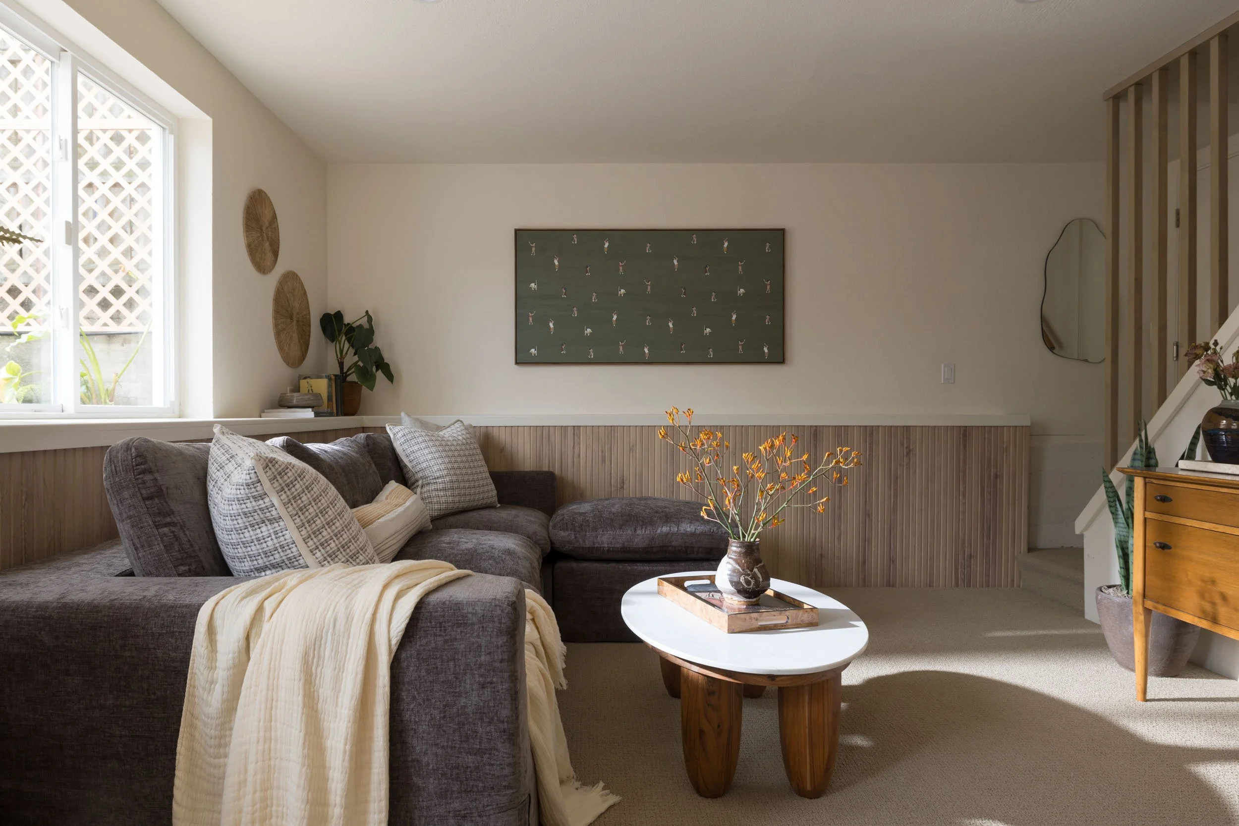 Living room with a brown sectional sofa, decorative pillows, a cream blanket, a wooden side table with a vase of orange flowers, a window with lattice, wall art, plants, and a staircase.