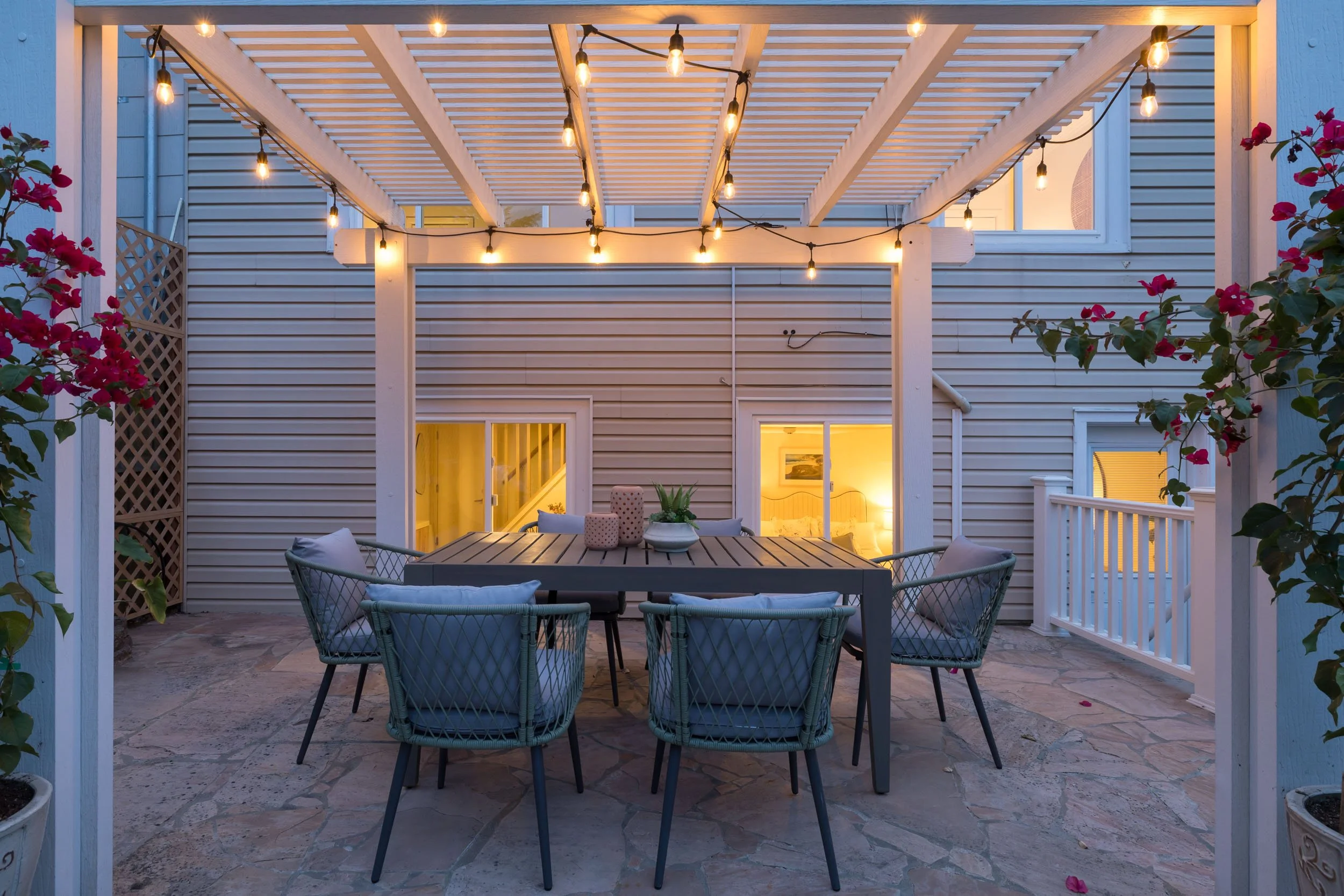 An outdoor patio with a rectangular table surrounded by six chairs, decorated with potted plants and candles. Overhead string lights illuminate the patio, which is enclosed by a white pergola. The background shows the house with lit windows and slidi