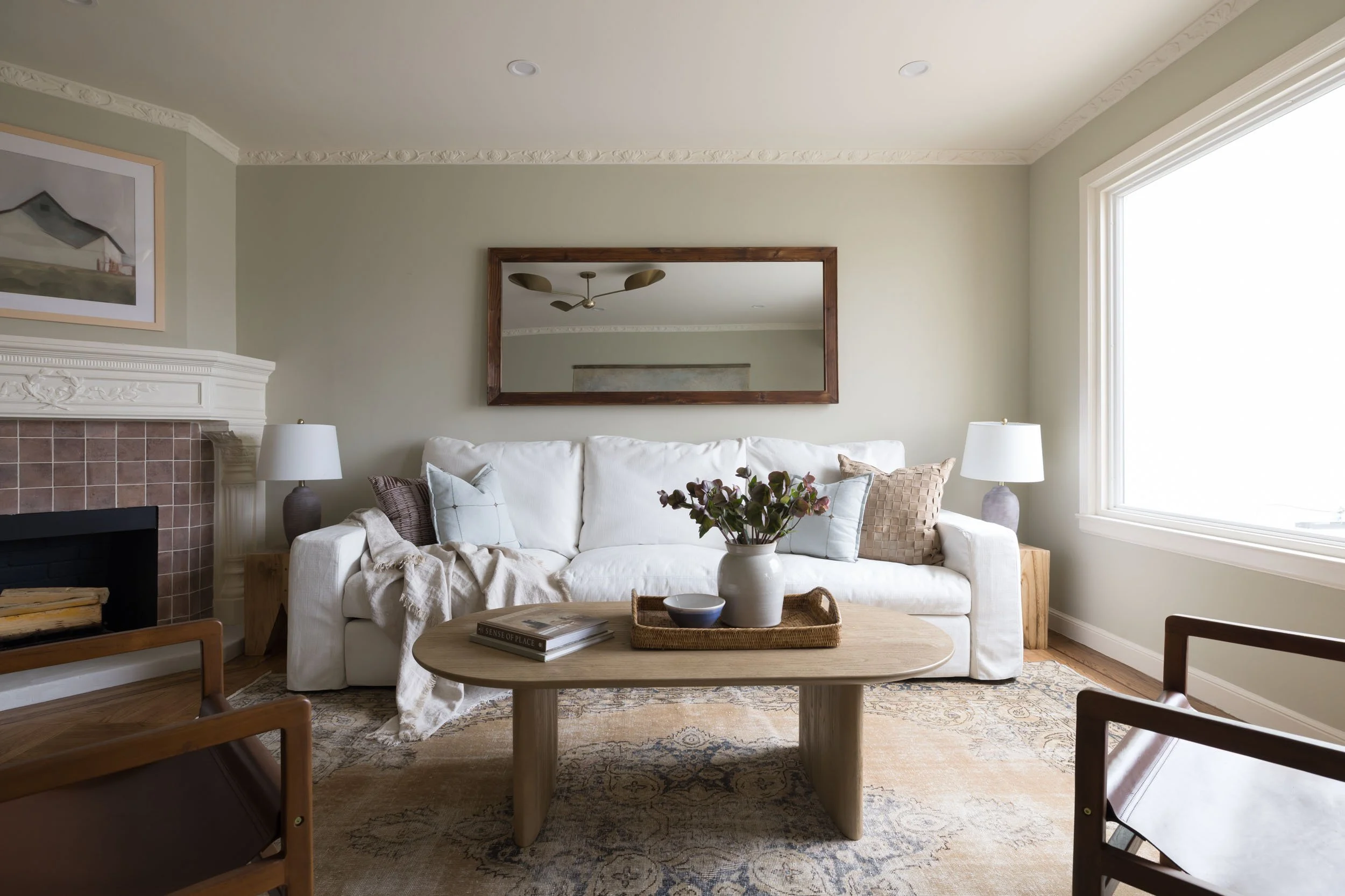 Bright living room with a white sofa, wooden coffee table with a vase of flowers, a mirror above the sofa, and large window letting in natural light.