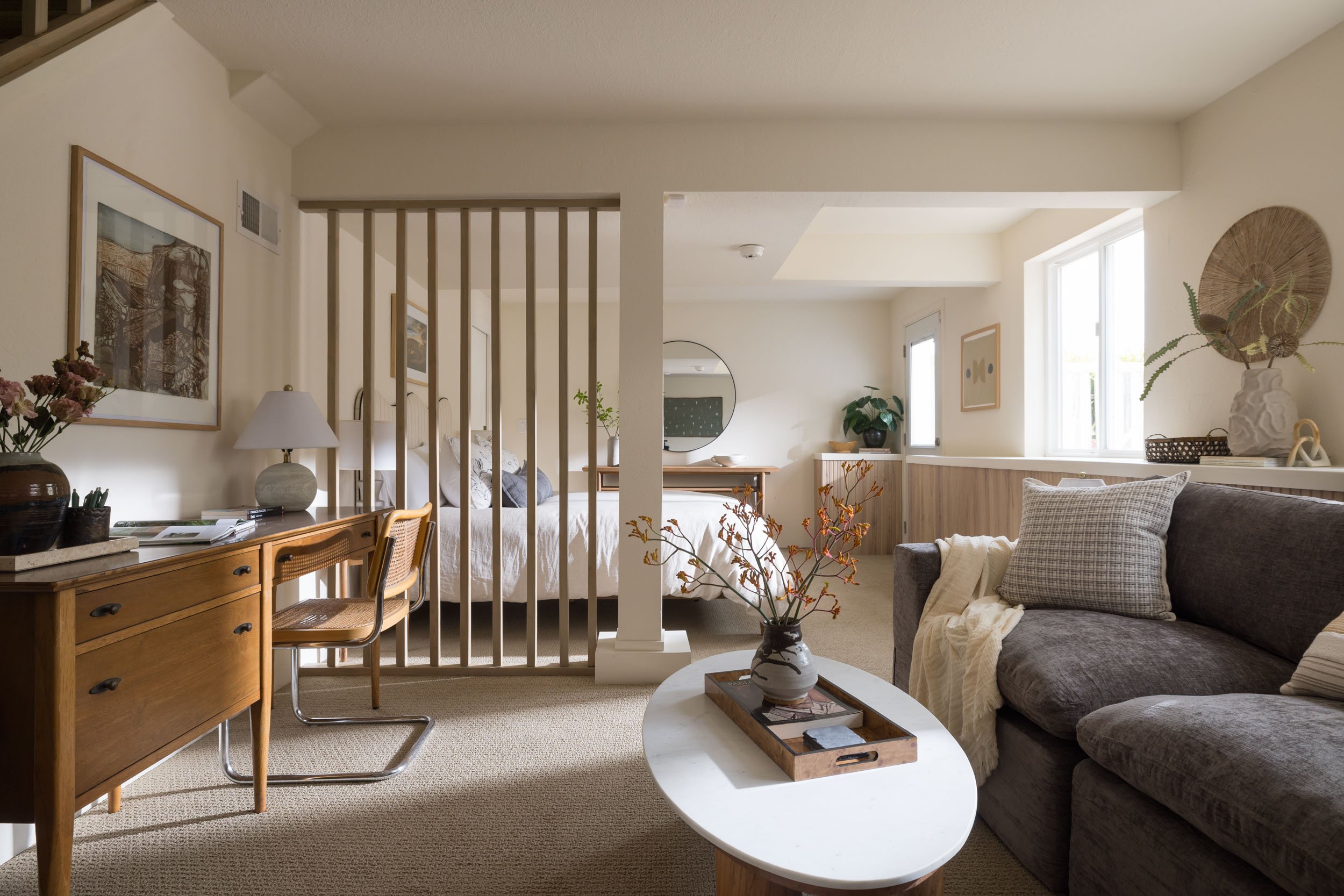 A cozy living space with a bedroom and living room area, featuring a wooden desk with a chair, a sofa with cushions, a coffee table with a vase of flowers, and large windows providing natural light.