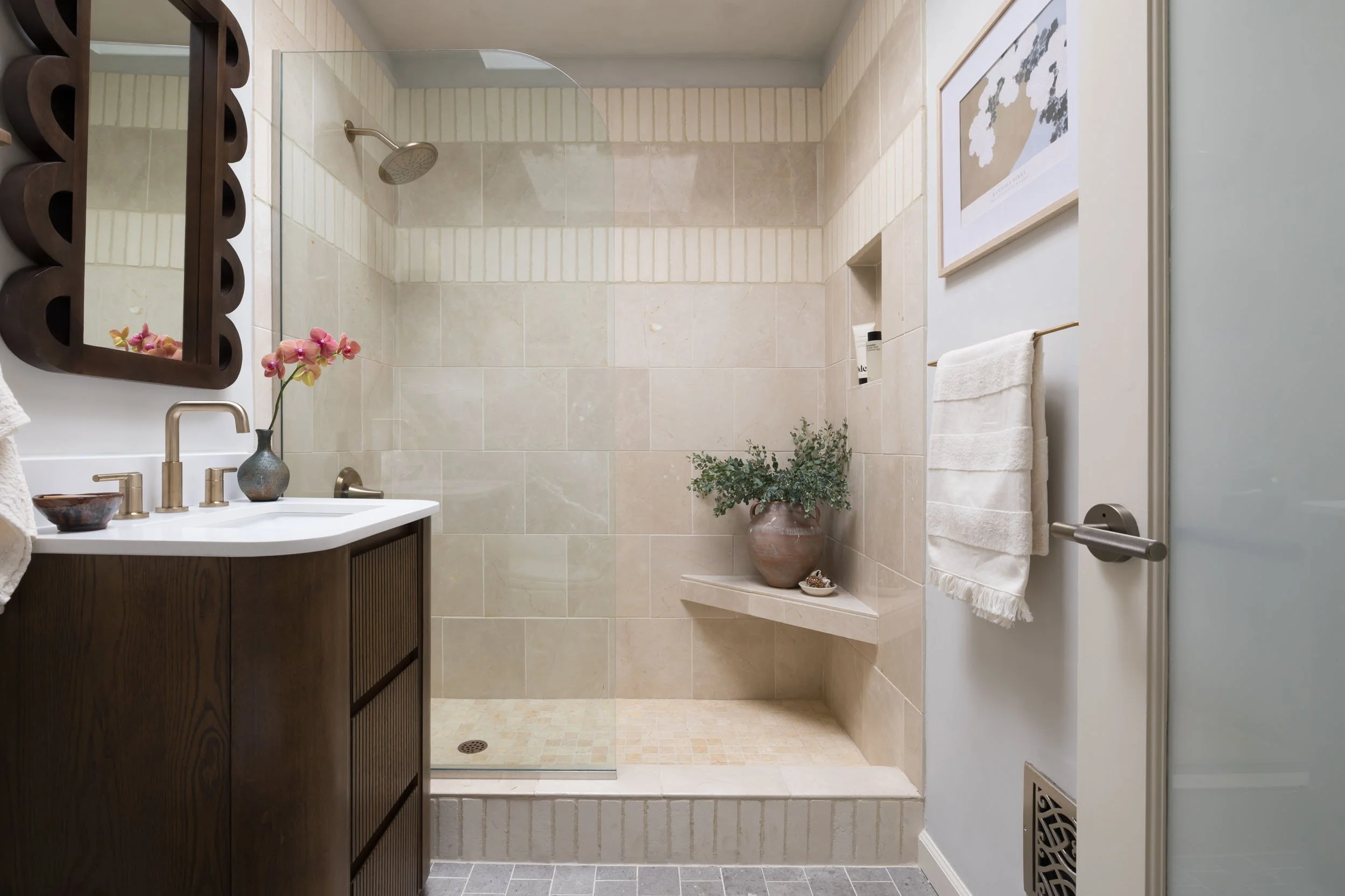 Bathroom with a shower area with beige tiles, a glass partition, a dark wood vanity with a white countertop, a neutral-toned decorative mirror, a vase with orchids, and a plant on a shelf.