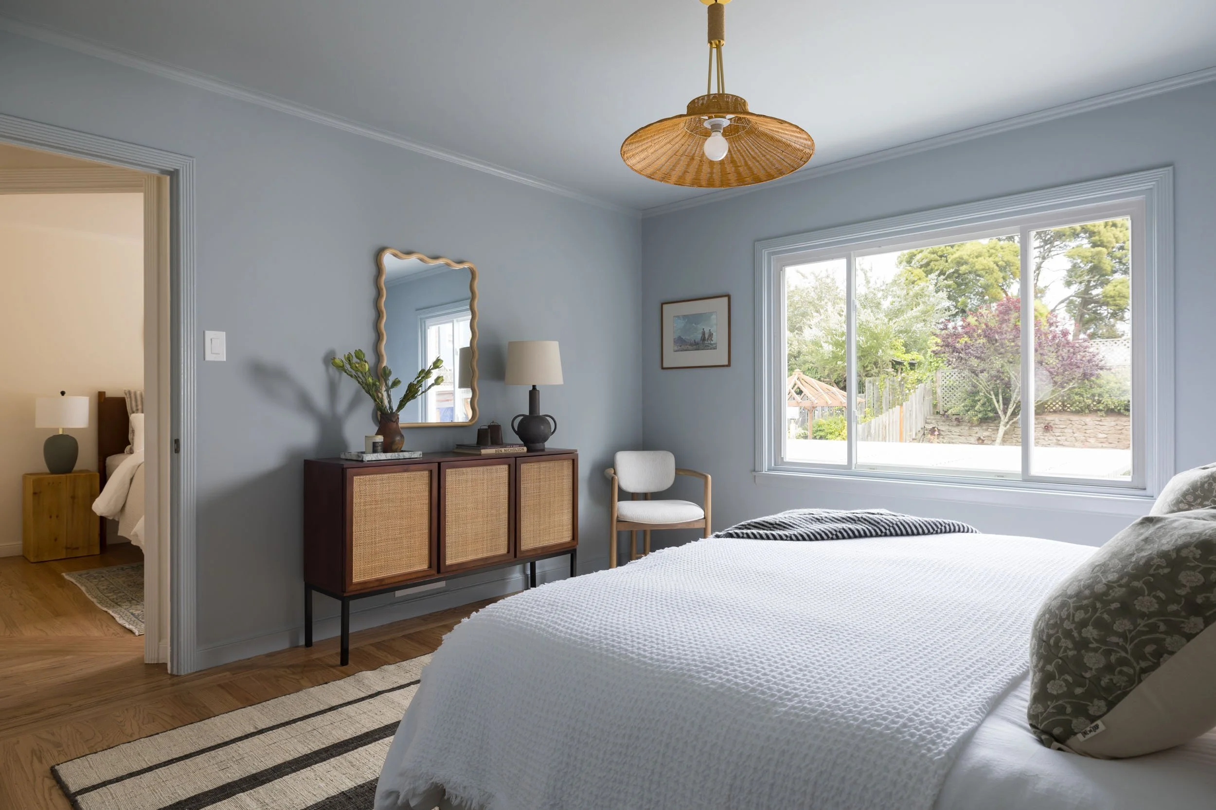 A cozy bedroom with a large window overlooking a garden, a white bed with patterned pillows, a wooden sideboard with a mirror and decor, and a doorway leading to a second bedroom.