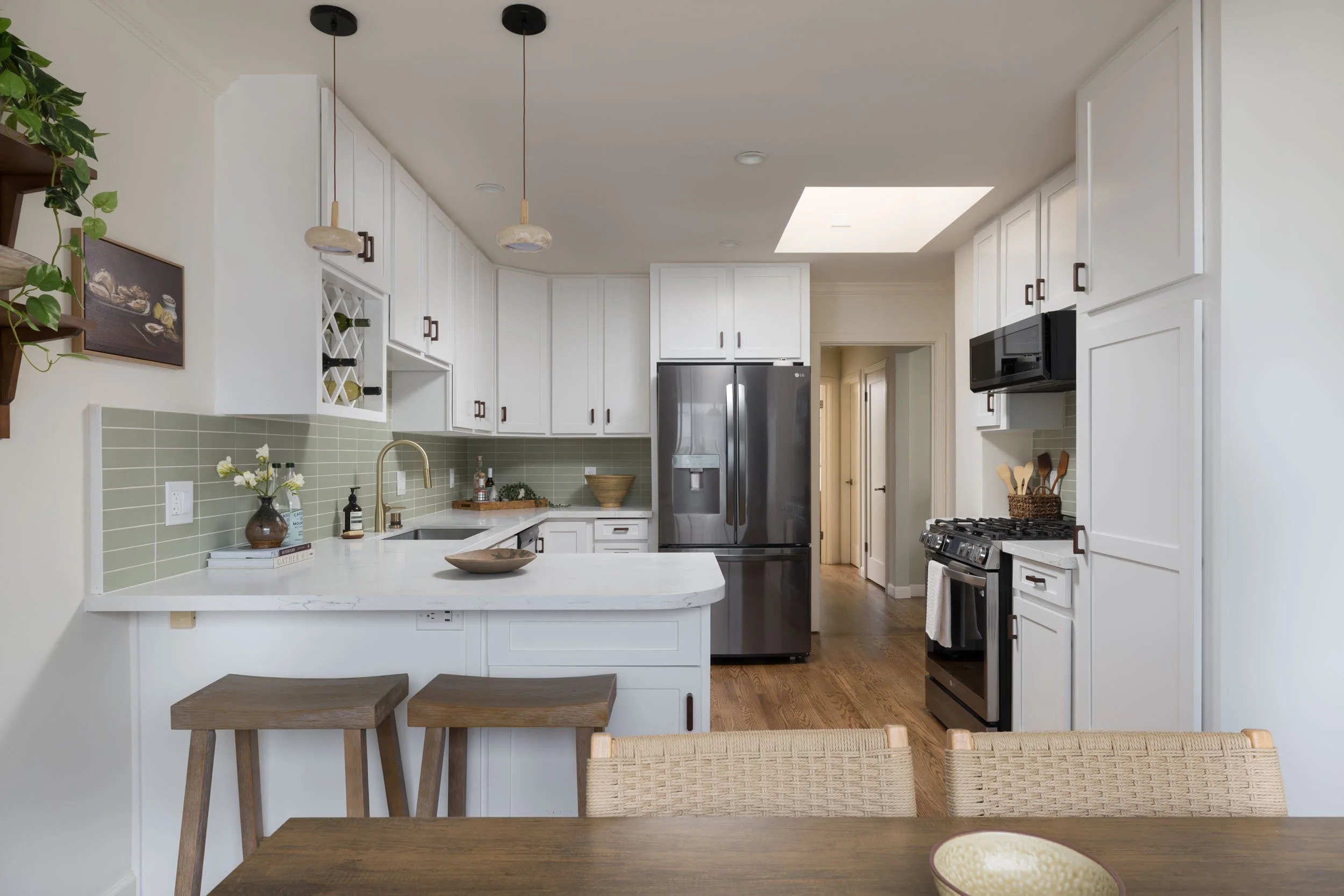Modern white kitchen with green tile backsplash, stainless steel refrigerator, black microwave, stove, and wood accents. There are two hanging pendant lights and a wooden dining table with woven chairs.