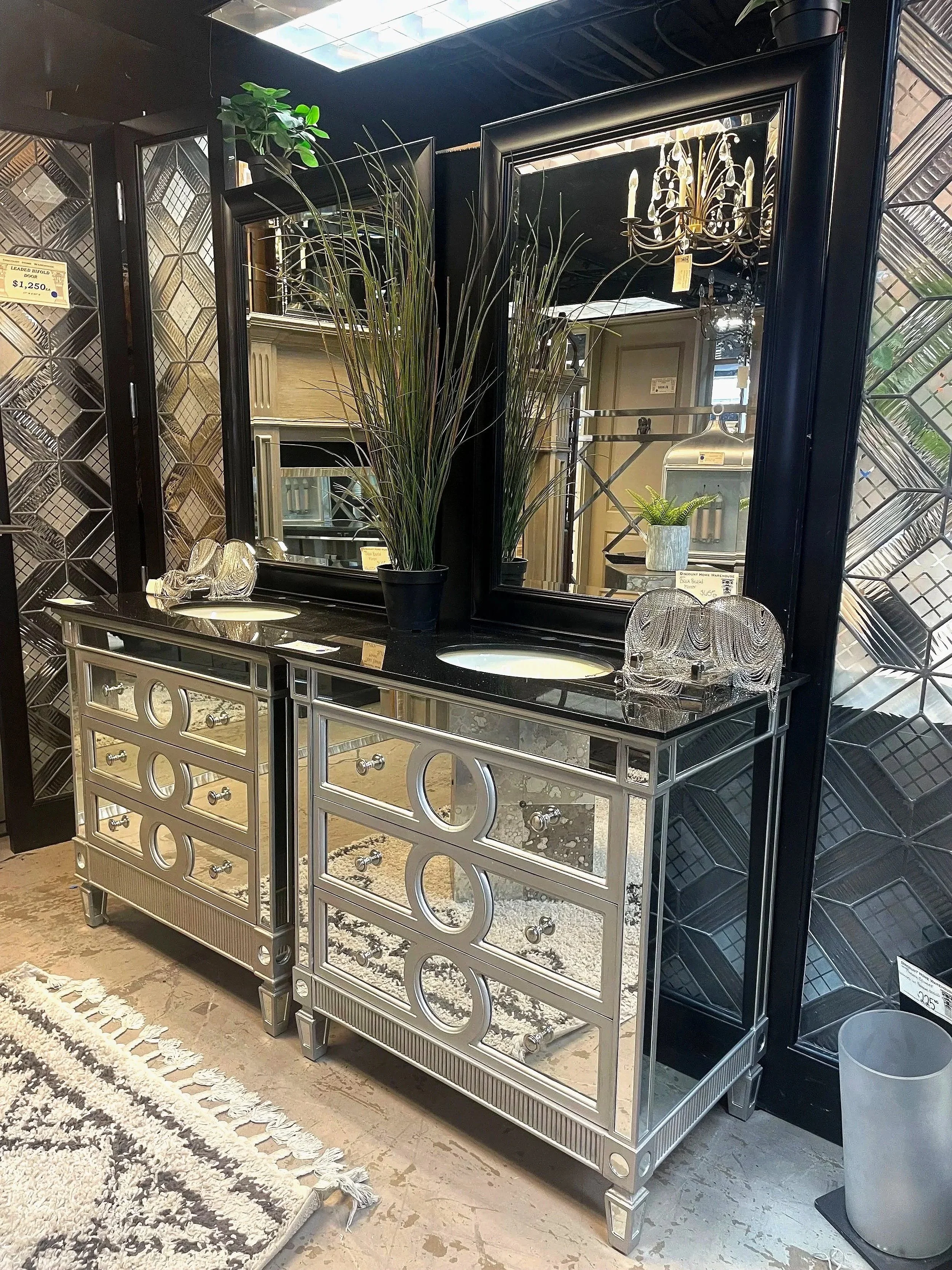 A mirrored dressing table with a silver frame, two sinks, and decorative knobs. Behind it, a large mirror reflects a chandelier and other furniture items in a home decor store.