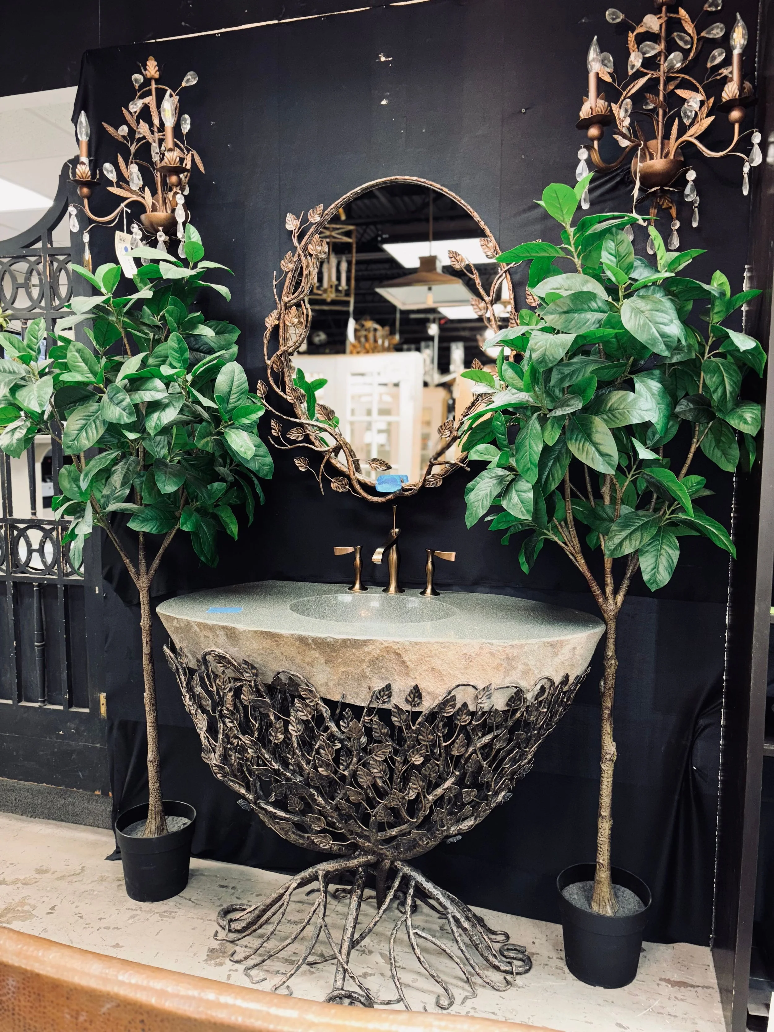 Decorative wall mirror with leaf design and two wall-mounted candle sconces with crystal accents, surrounded by two potted ficus trees.