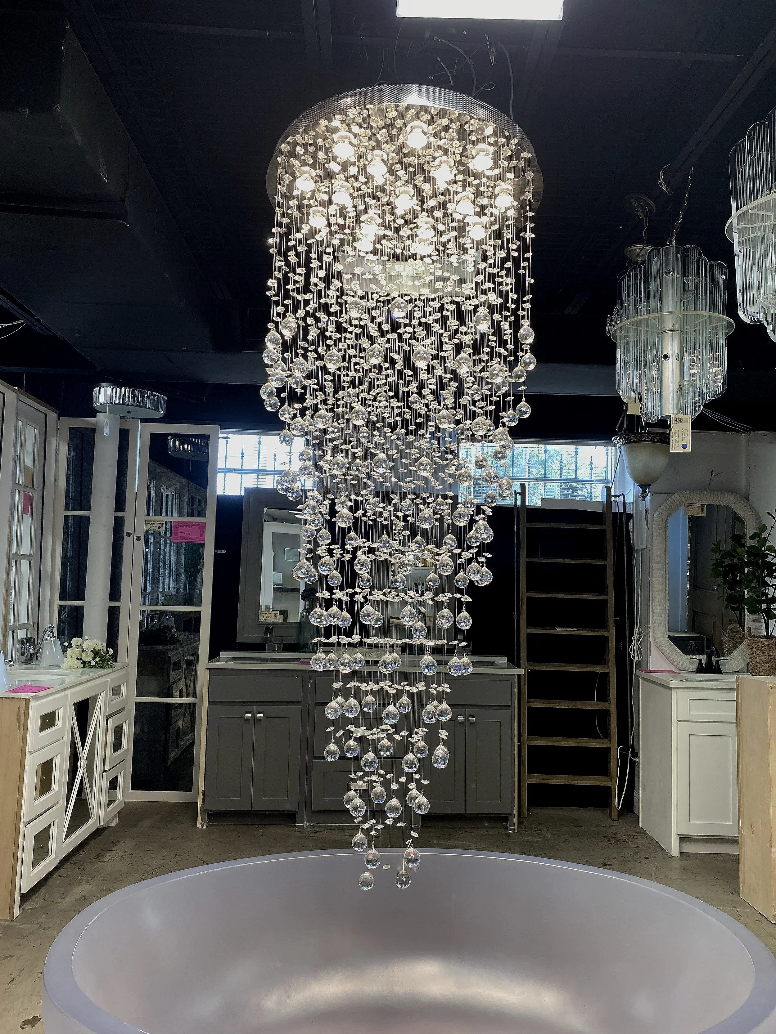 A store display featuring a large, modern crystal chandelier hanging above a circular table with a gray surface. Surrounding the chandelier are other lighting fixtures and furniture items.