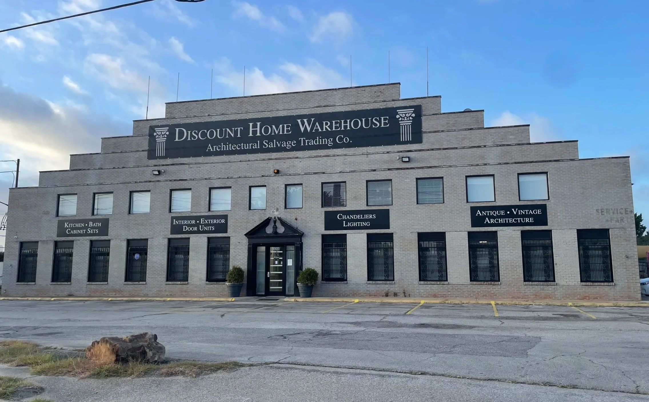 Front view of a commercial building with a sign reading 'Discount Home Warehouse' and smaller signs advertising kitchen and bath cabinets, interior and exterior door units, chandeliers and lighting, and antique and vintage architecture. The building has a stepped design, multiple windows with black bars, and two potted plants at the entrance.